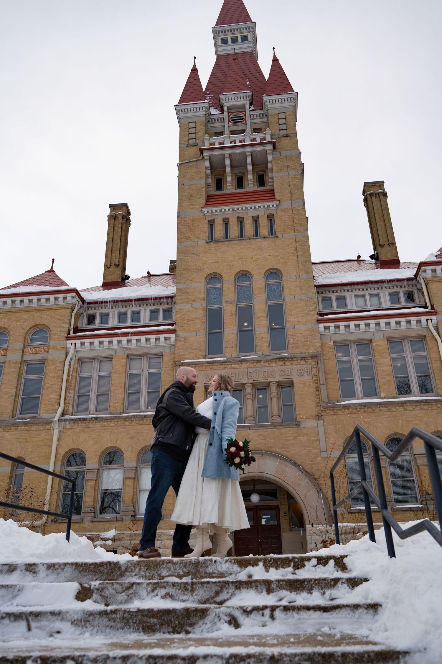 Couple standing on snowy steps of a historic brick building in winter wedding attire.