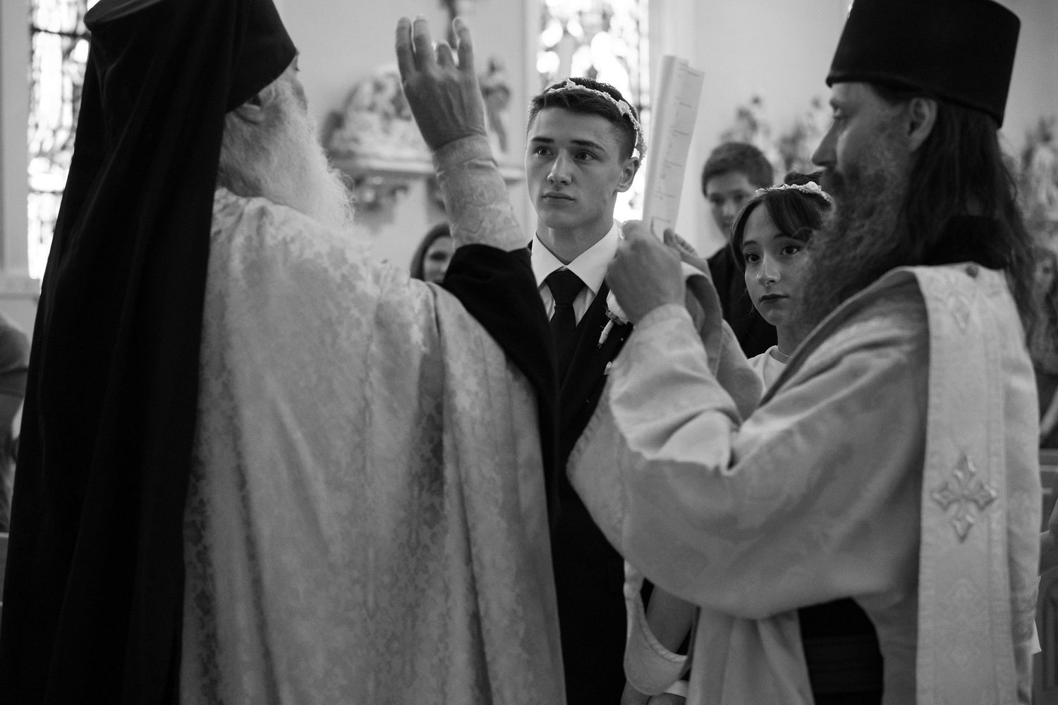 Black and white photo of a groom receiving a blessing from Orthodox priests during wedding.