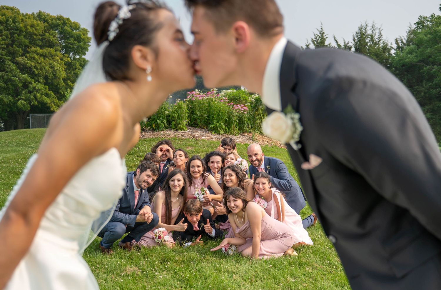 Bride and groom kiss in foreground while wedding party poses and cheers on grass behind them.