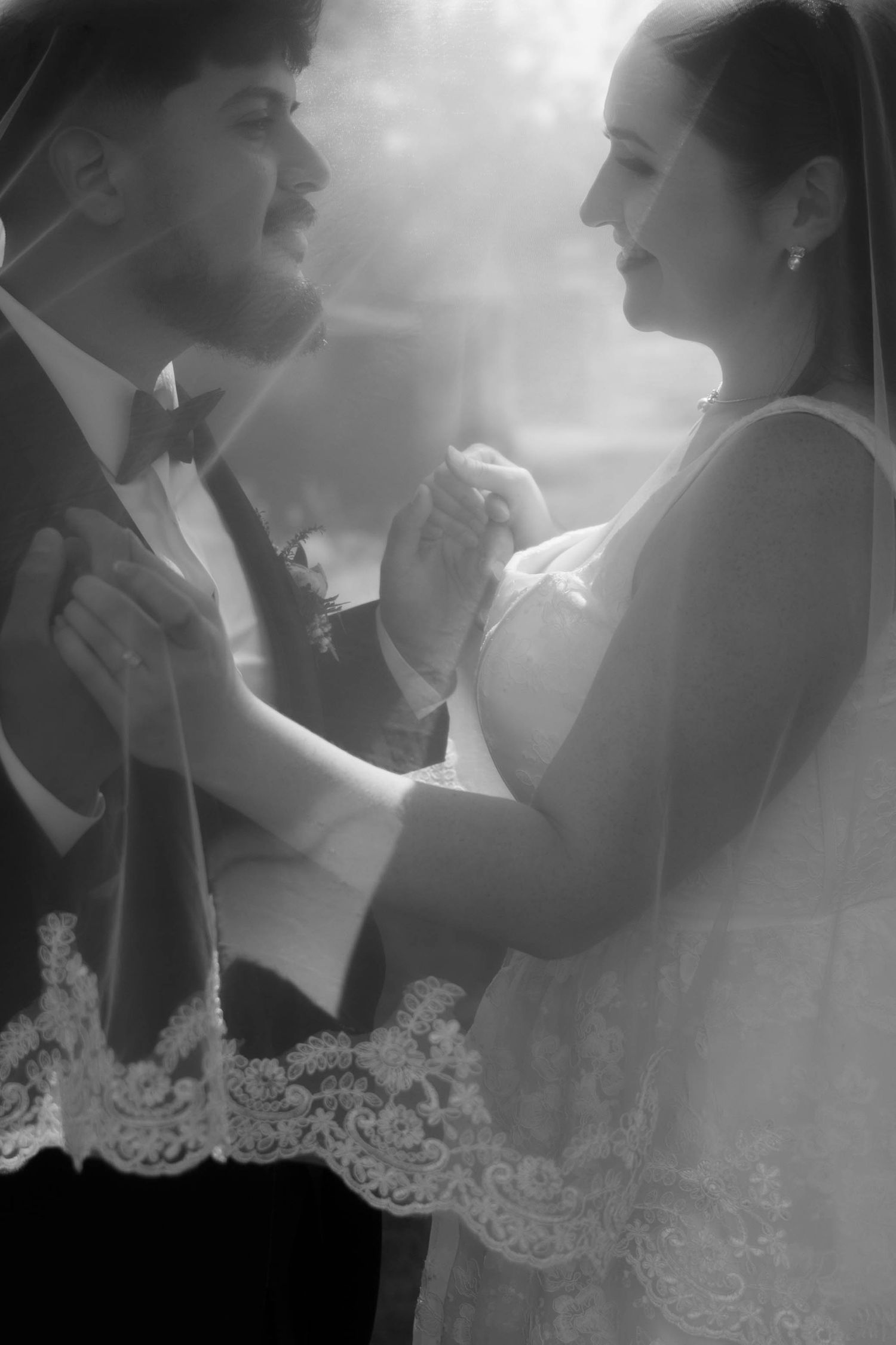 Black and white photo of a bride and groom dancing together, smiling, under a lace veil with soft bokeh lighting.