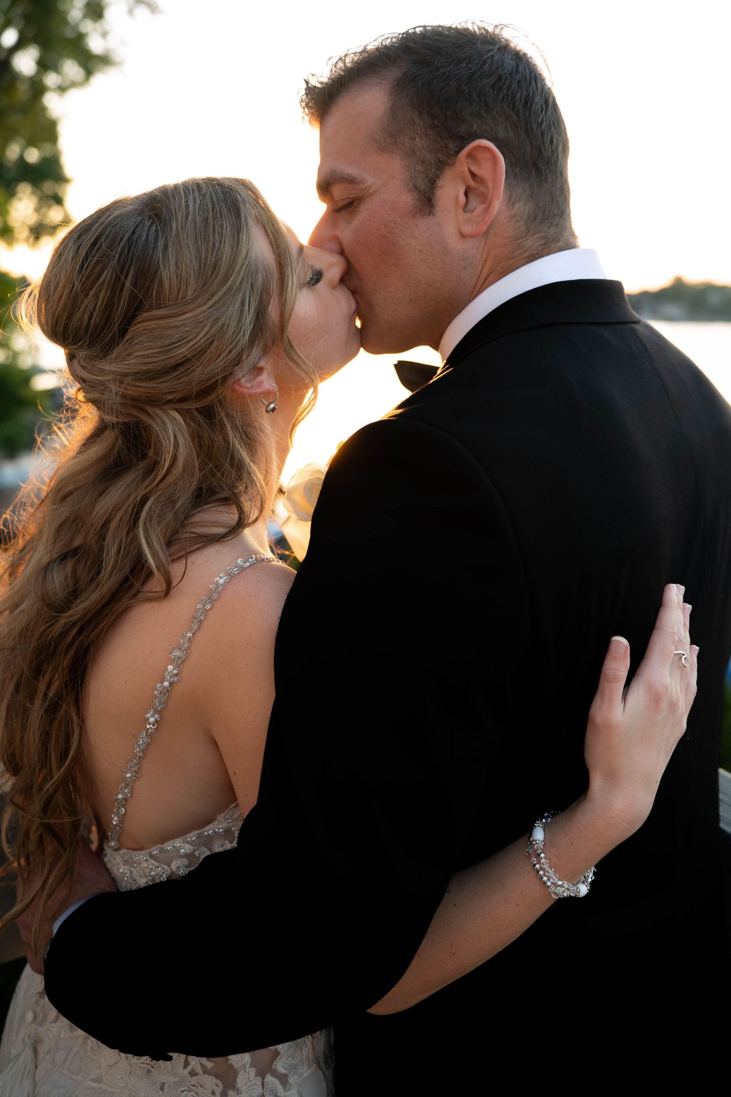A bride and groom share a romantic kiss at sunset, with the bride in a backless gown and groom in a black tuxedo.
