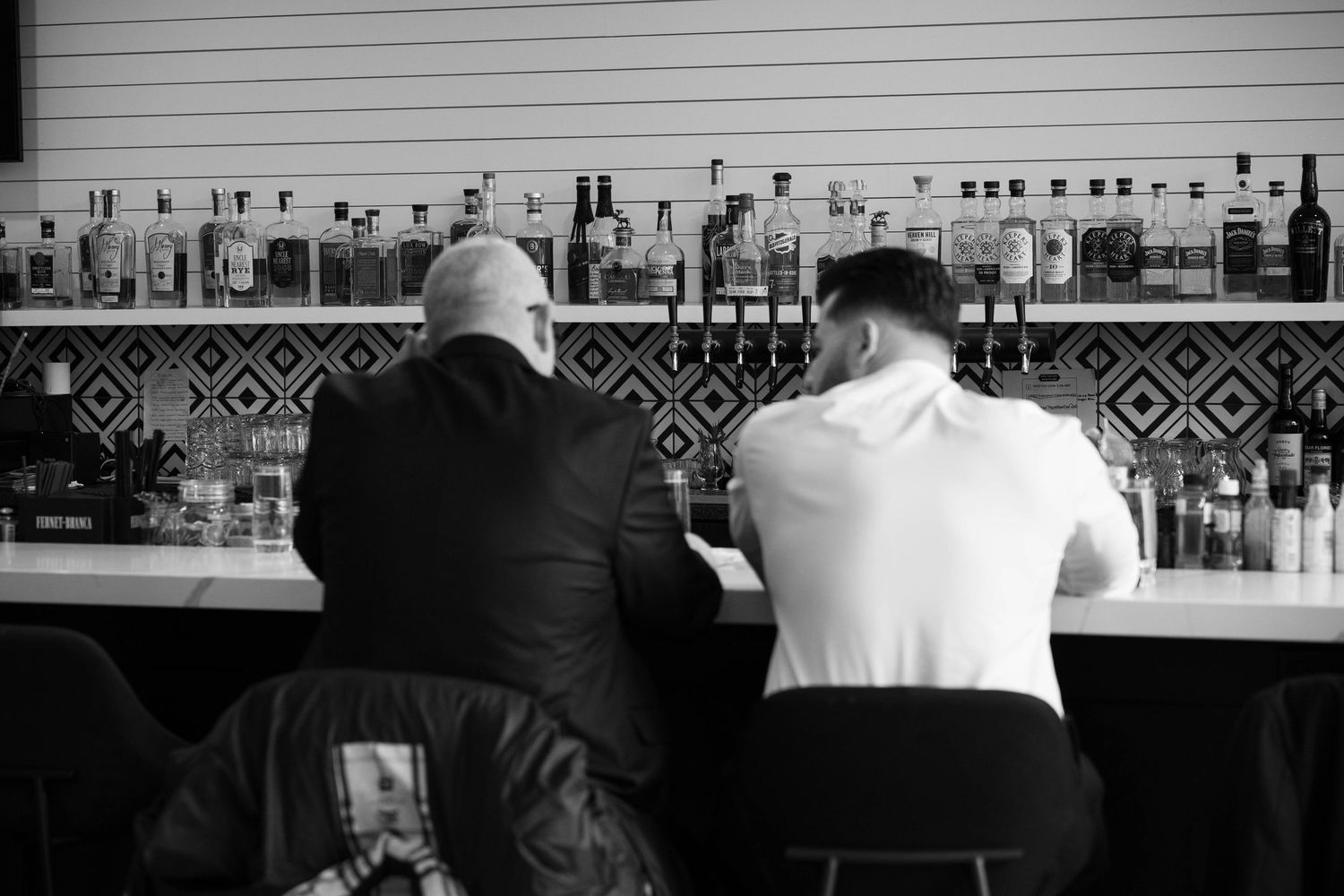 Two men sit at a well-stocked bar in a black and white candid wedding reception photo.