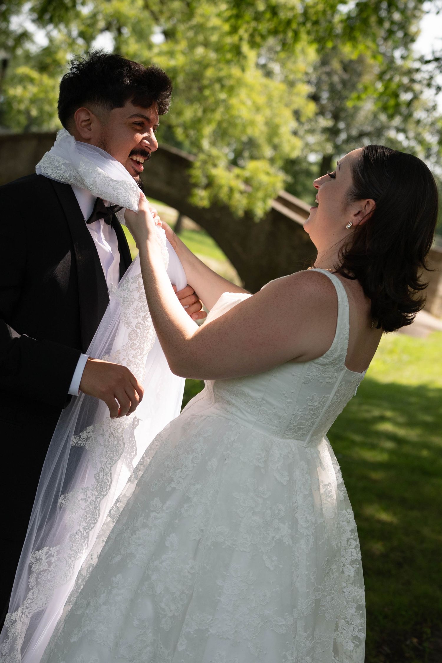 Laughing bride playfully wraps her veil around groom's neck as couple shares a joyful moment in green park.