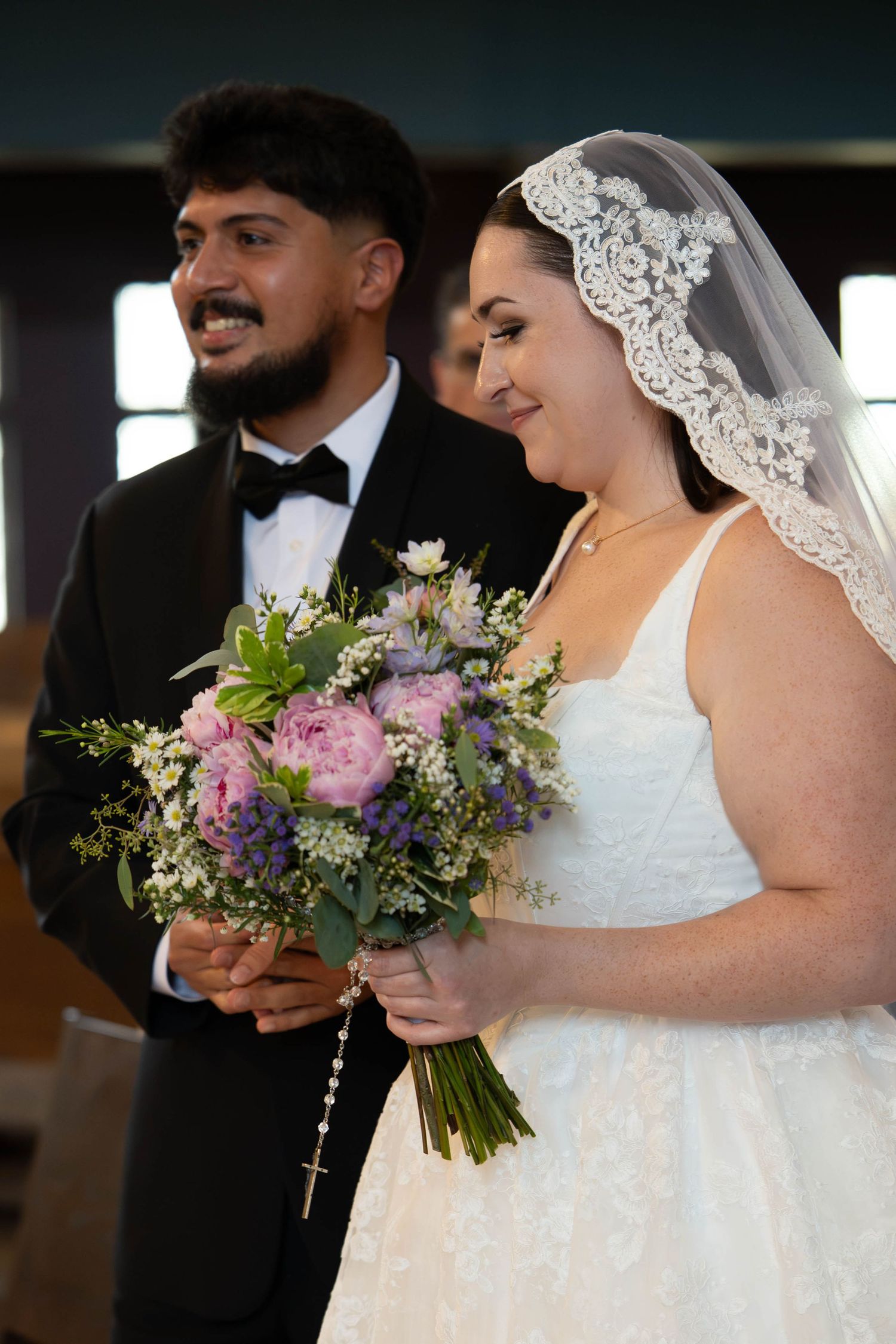 Bride holding purple and pink floral bouquet with rosary stands beside smiling groom in church.
