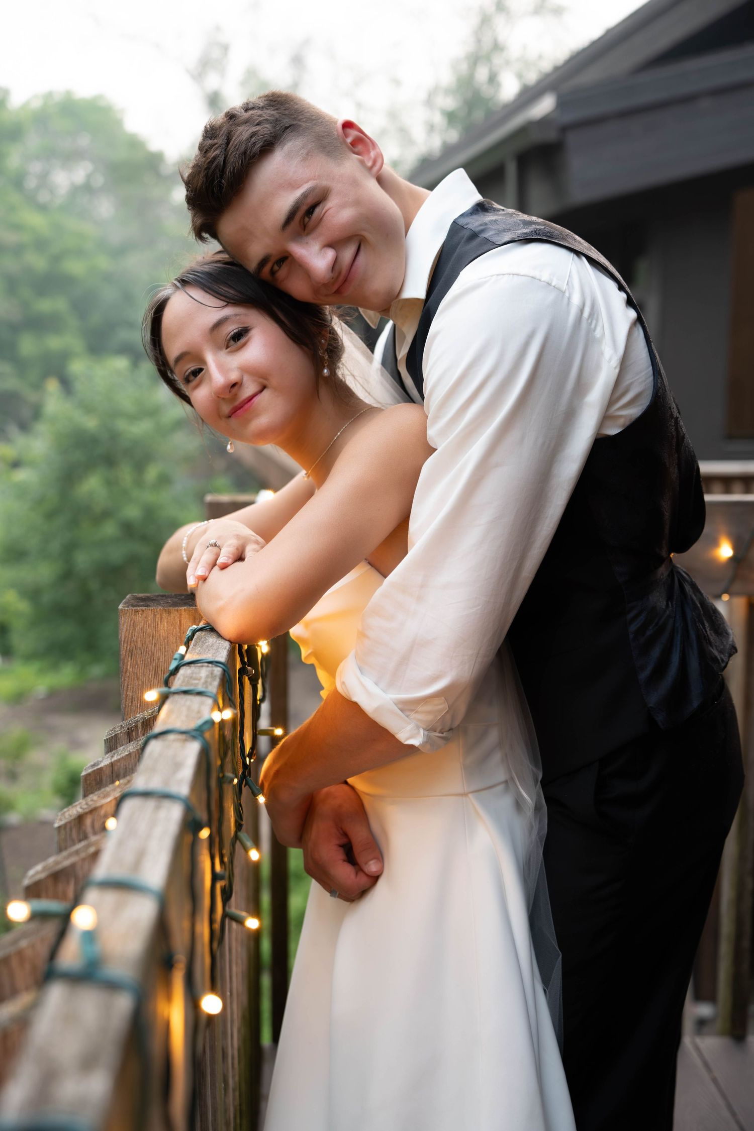 Happy newlywed couple embracing on decorated balcony with string lights at dusk.