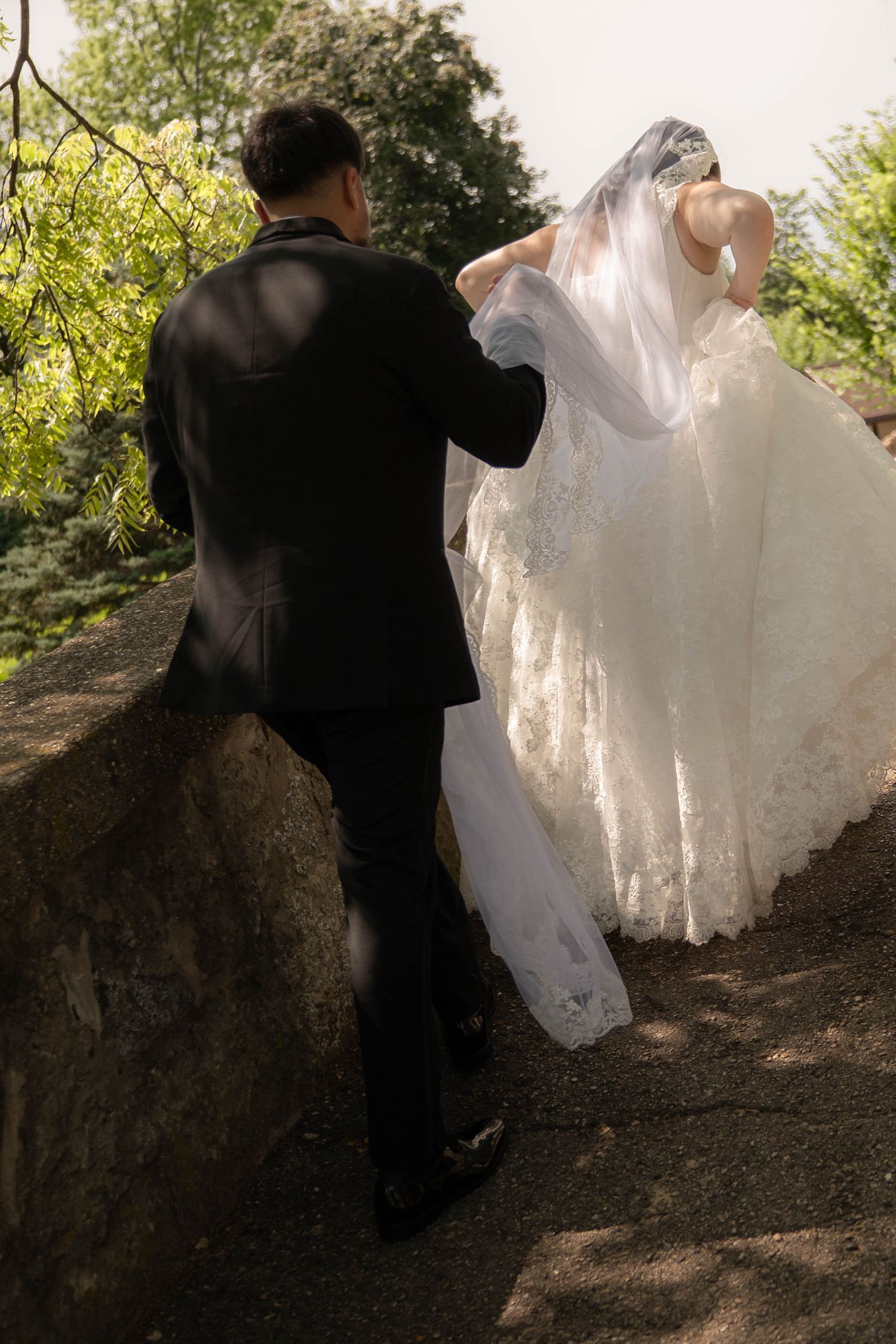 Groom in black suit helps bride in white lace gown with her veil while walking along a stone path outdoors.