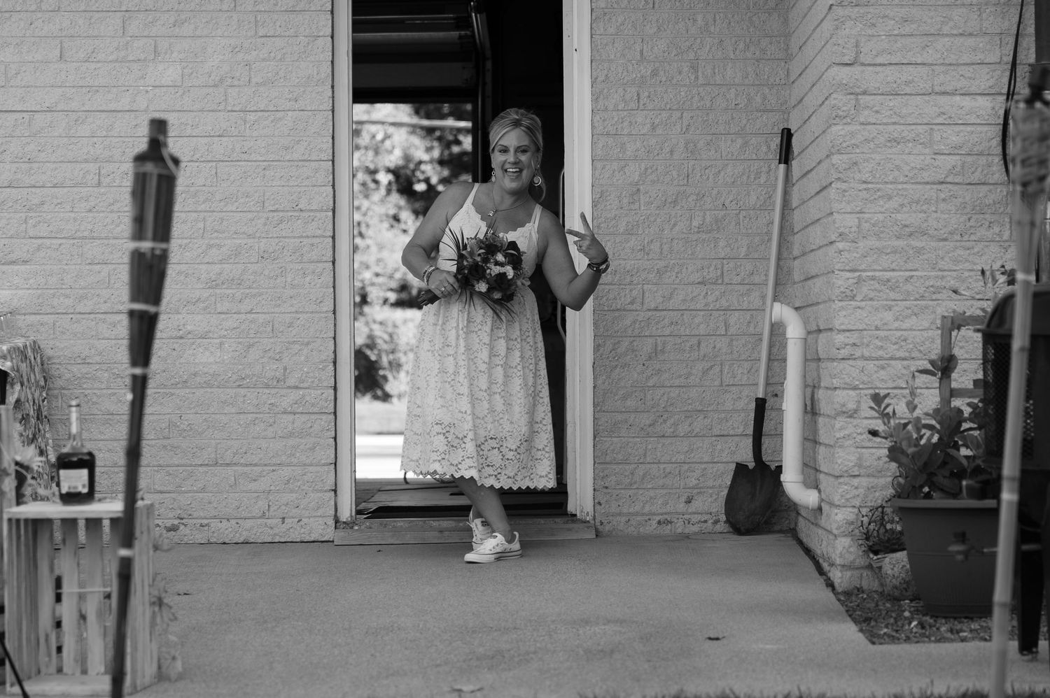 Bride in white lace dress holding bouquet, walking through doorway in black and white photo.