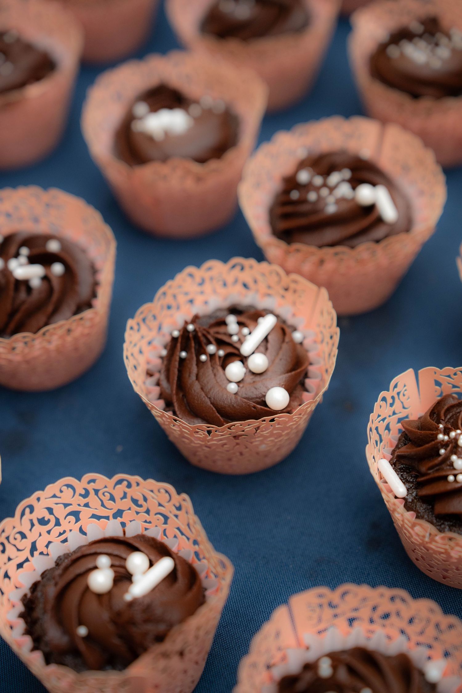 Chocolate cupcakes with white pearl sprinkles in decorative lace pink wrappers on blue surface.