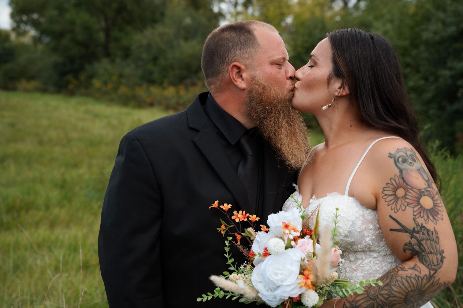Bride and groom sharing a kiss in a green meadow, bride holding colorful bouquet.