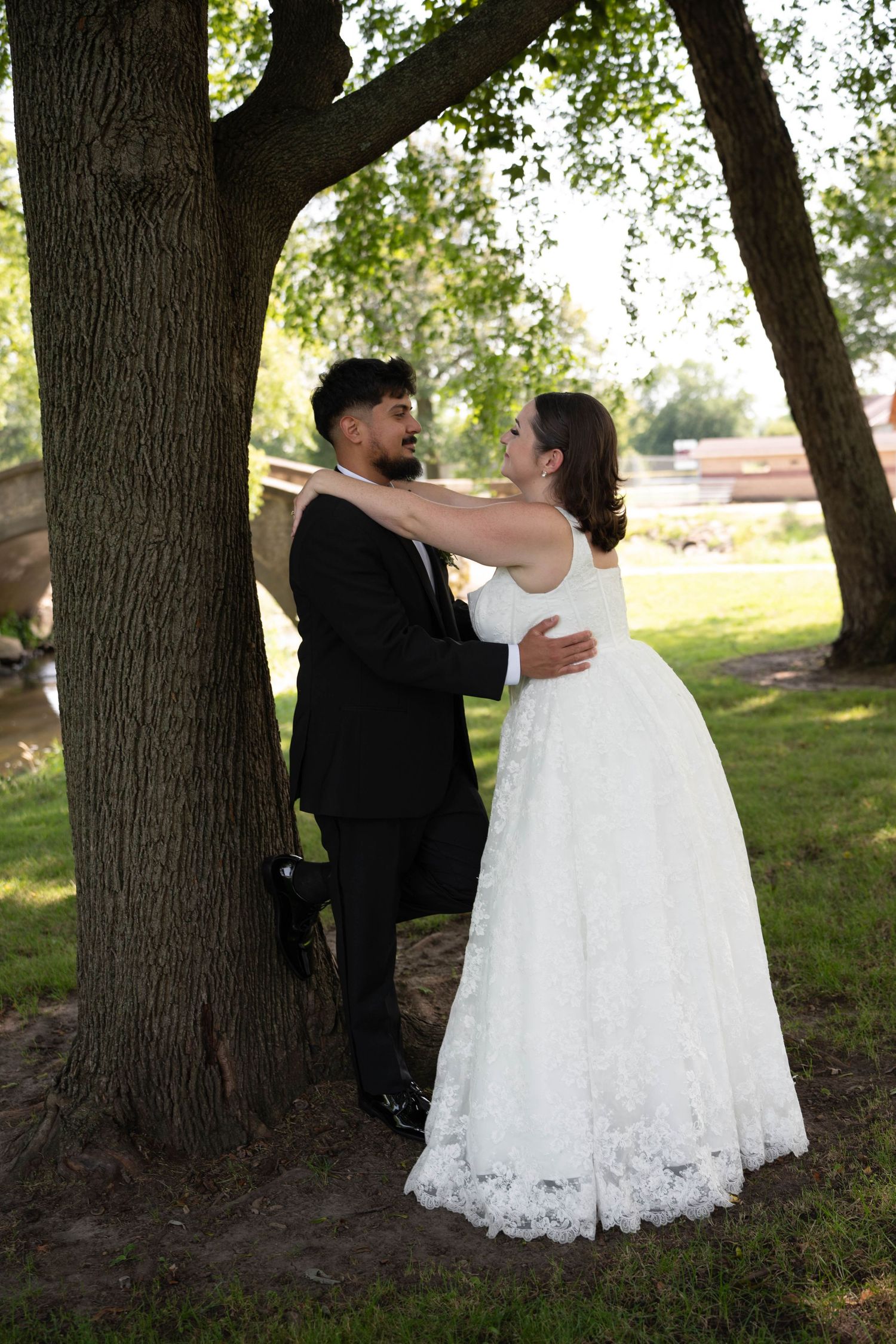 Bride in white lace gown and groom in black suit embrace romantically against a large tree in a park.