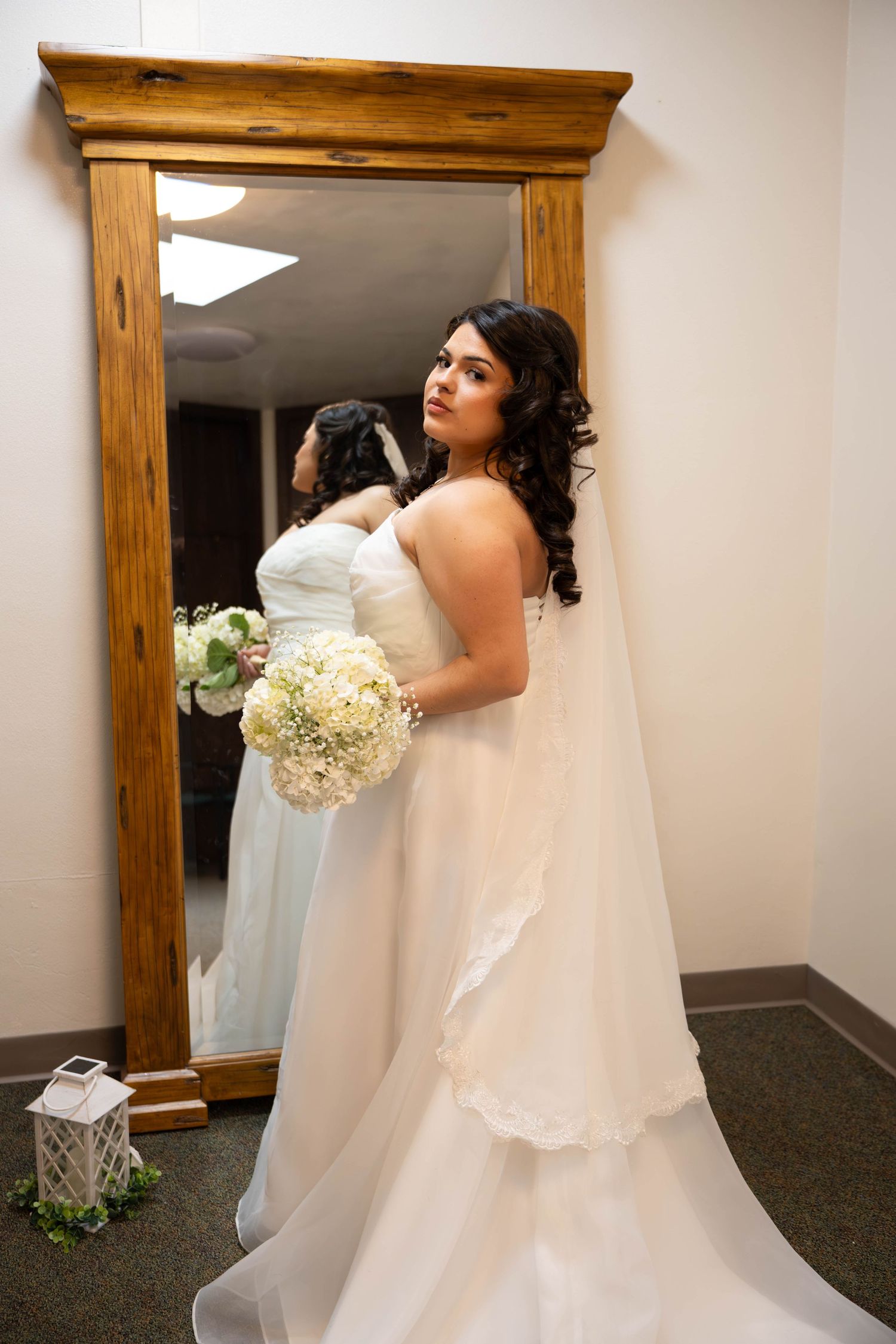 Elegant bride in strapless white gown holds bouquet while posing beside large wooden mirror.