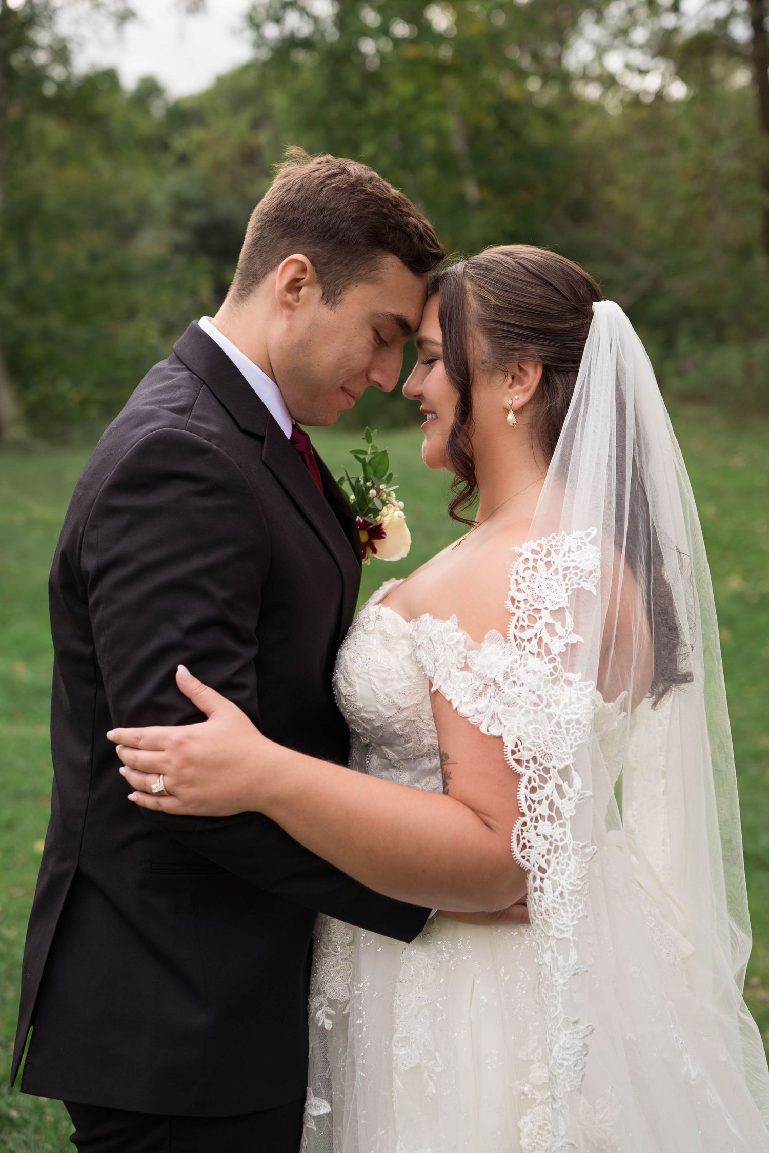 A bride and groom share a romantic forehead touch outdoors, she in lace gown with veil, he in black suit with burgundy boutonniere.