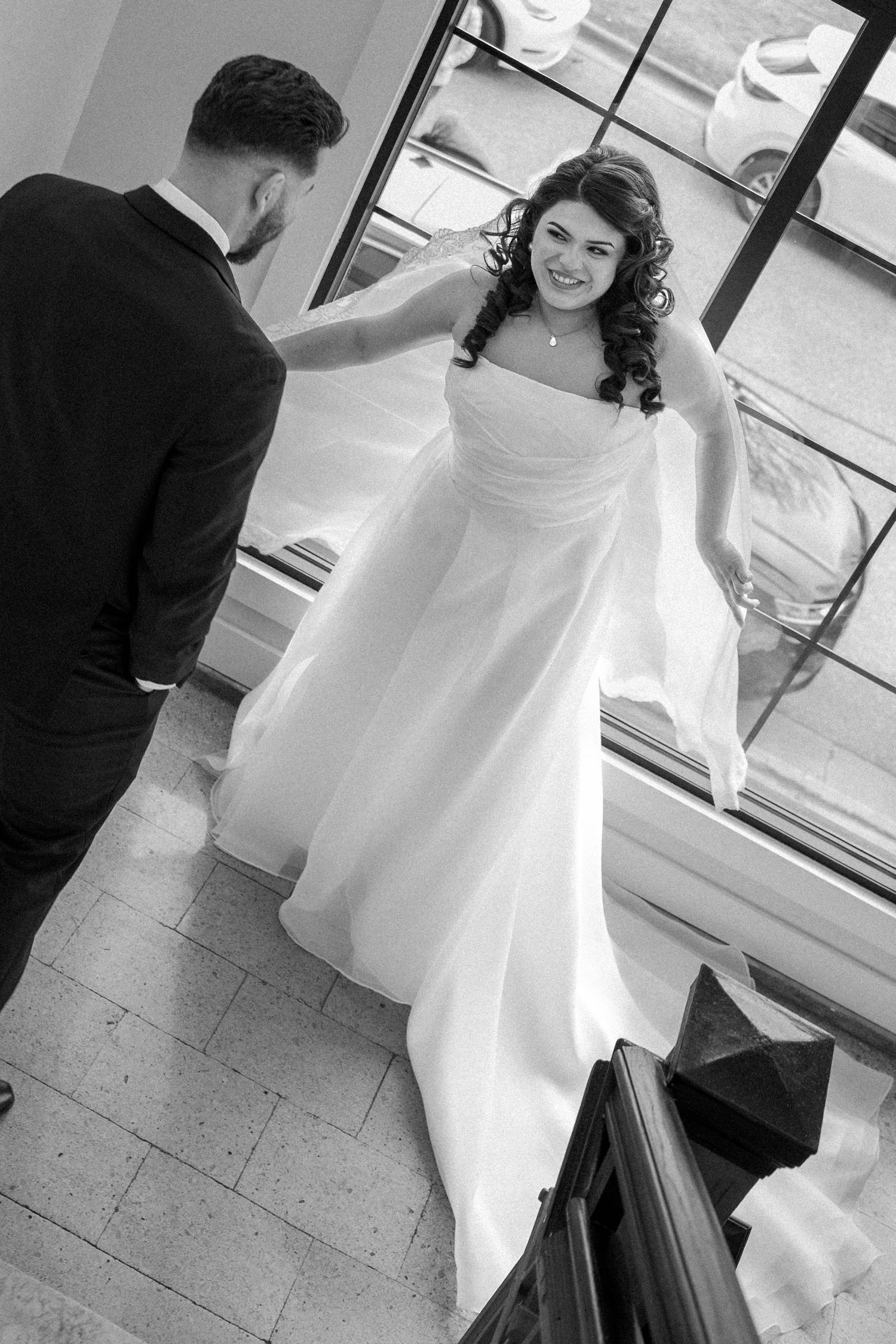 Bride in white gown smiles at groom on staircase in black and white wedding photo.