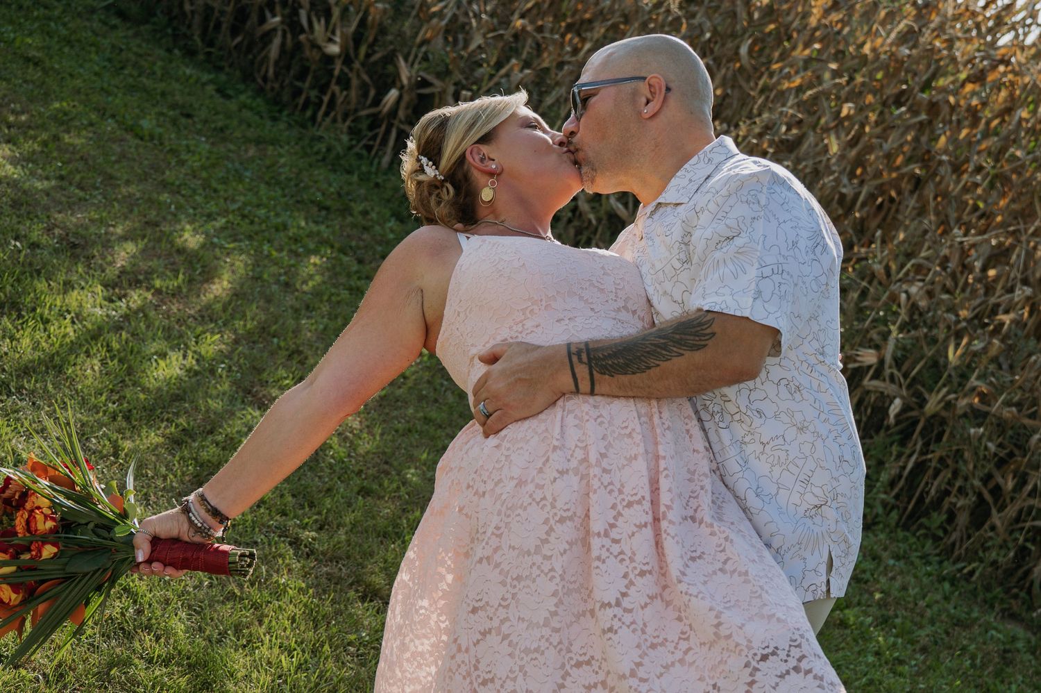 Wedding couple sharing a kiss near cornfield, bride in pink lace dress holding autumn bouquet.