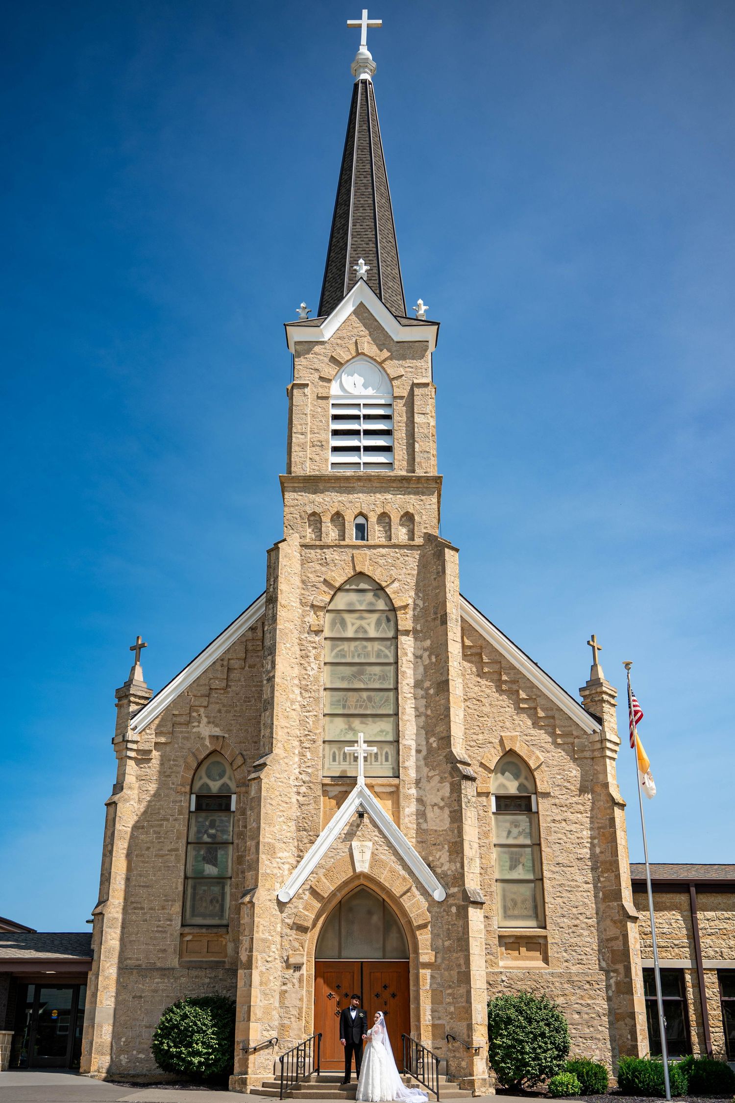 Bride and groom pose before historic stone Catholic church with tall steeple under blue sky.
