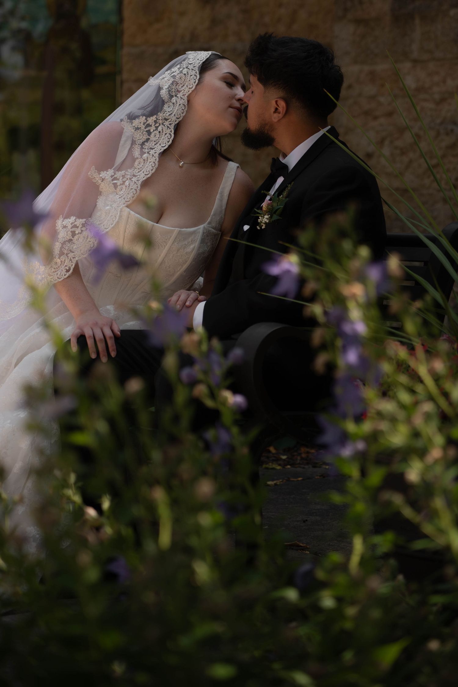 Bride and groom share a kiss while seated among purple wildflowers in a lush garden setting.