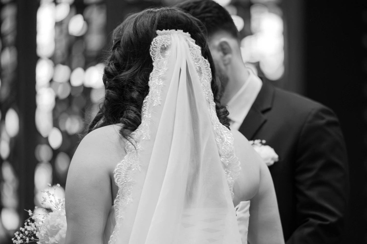 Black and white photo of bride and groom sharing first kiss, bridal veil draped over couple during ceremony.
