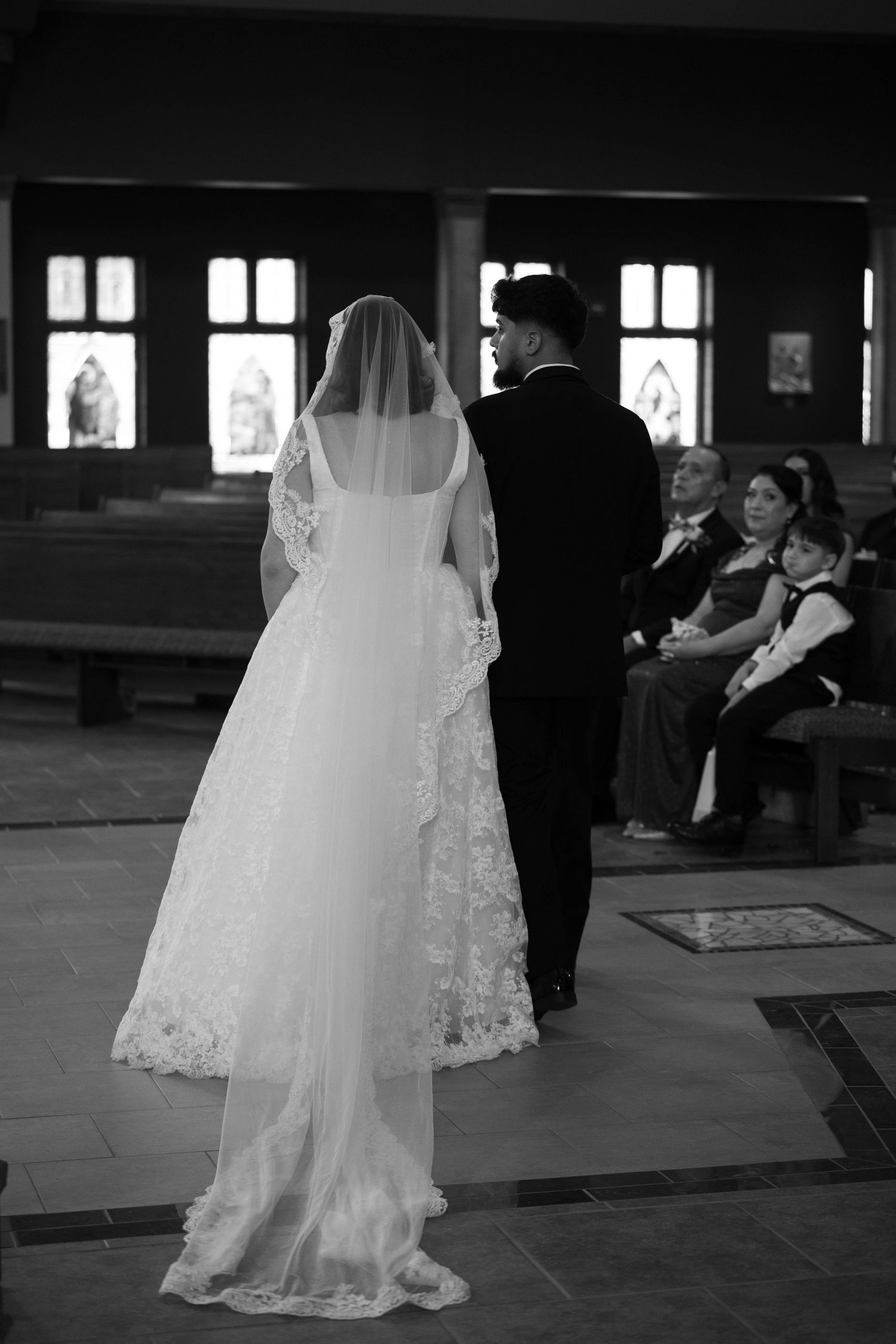 Black and white photo of bride in lace veil and groom standing at the altar with seated wedding guests.