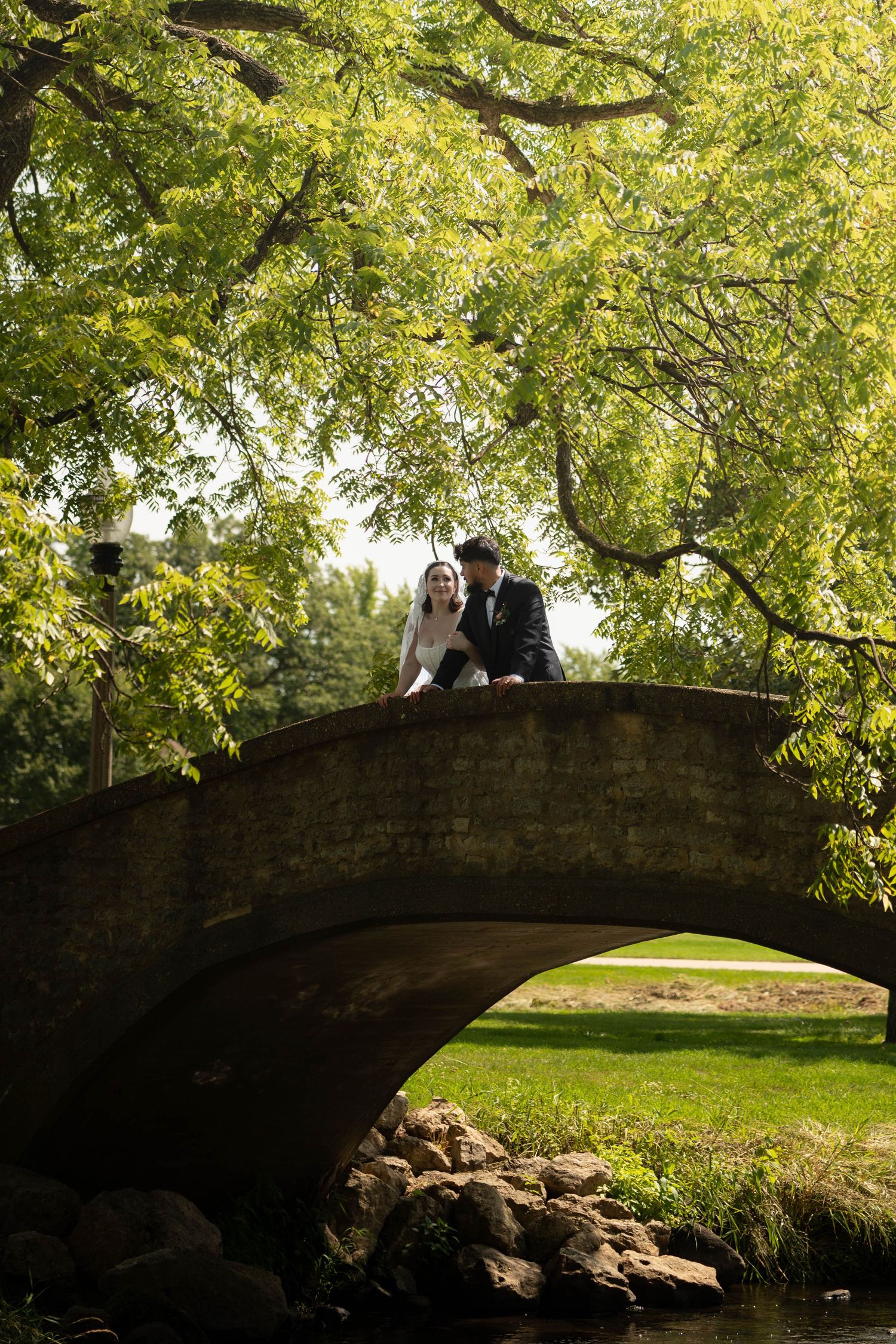 Bride and groom share a romantic moment on a stone arch bridge surrounded by lush green trees in a park.