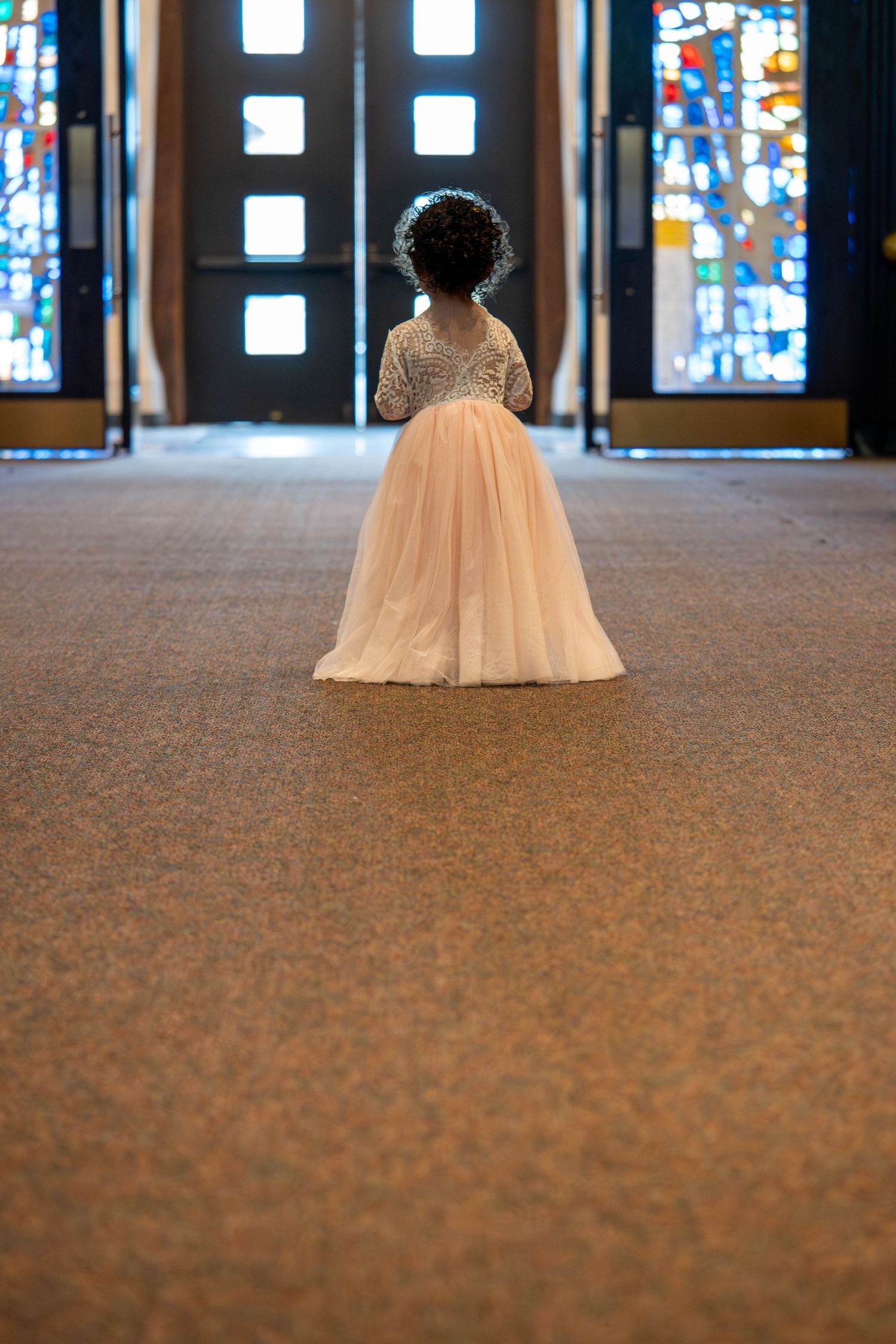 Adorable flower girl in white tulle dress walking alone down church aisle toward stained glass entrance doors.