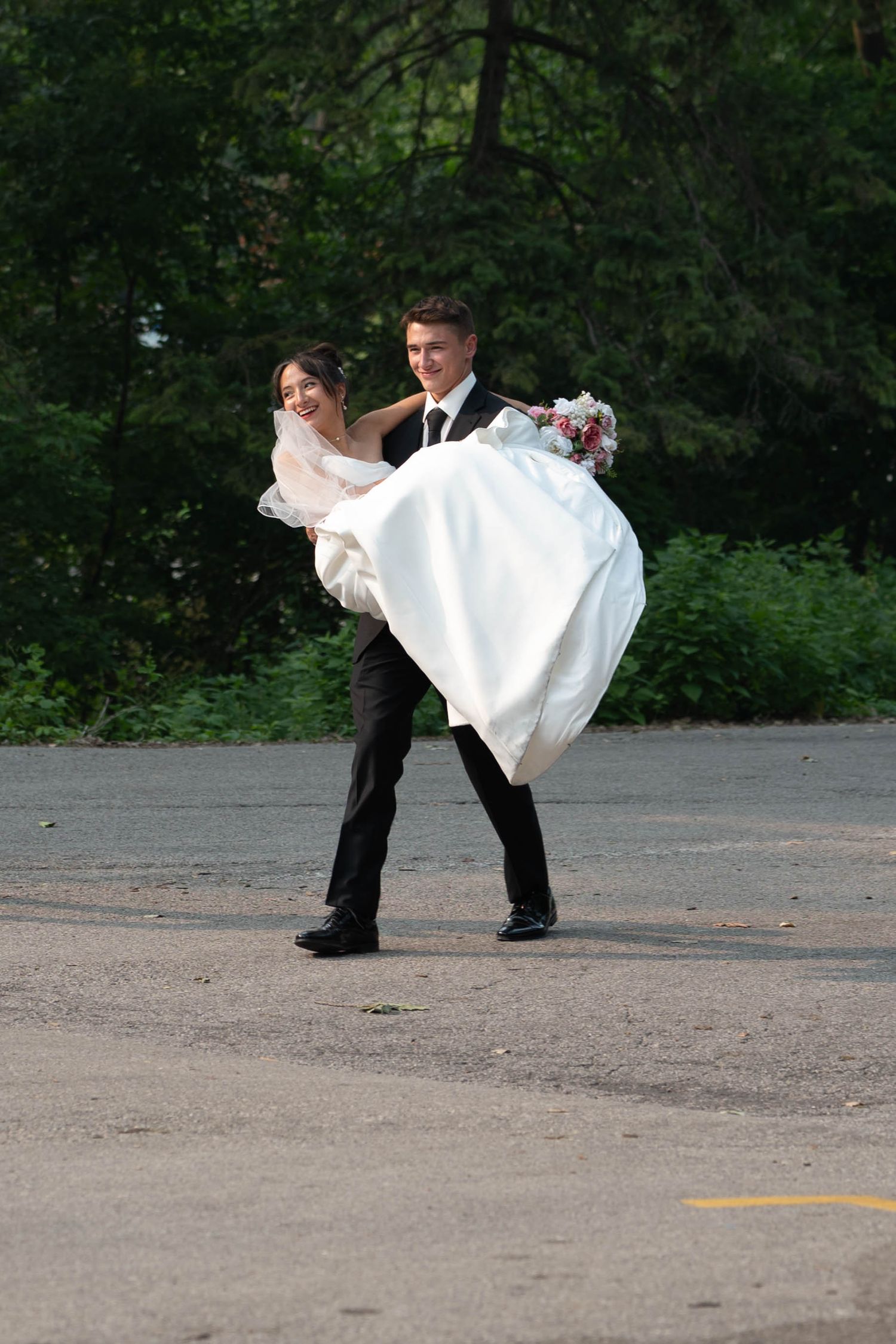 Smiling groom carries bride in white wedding gown outdoors near green trees on pavement.