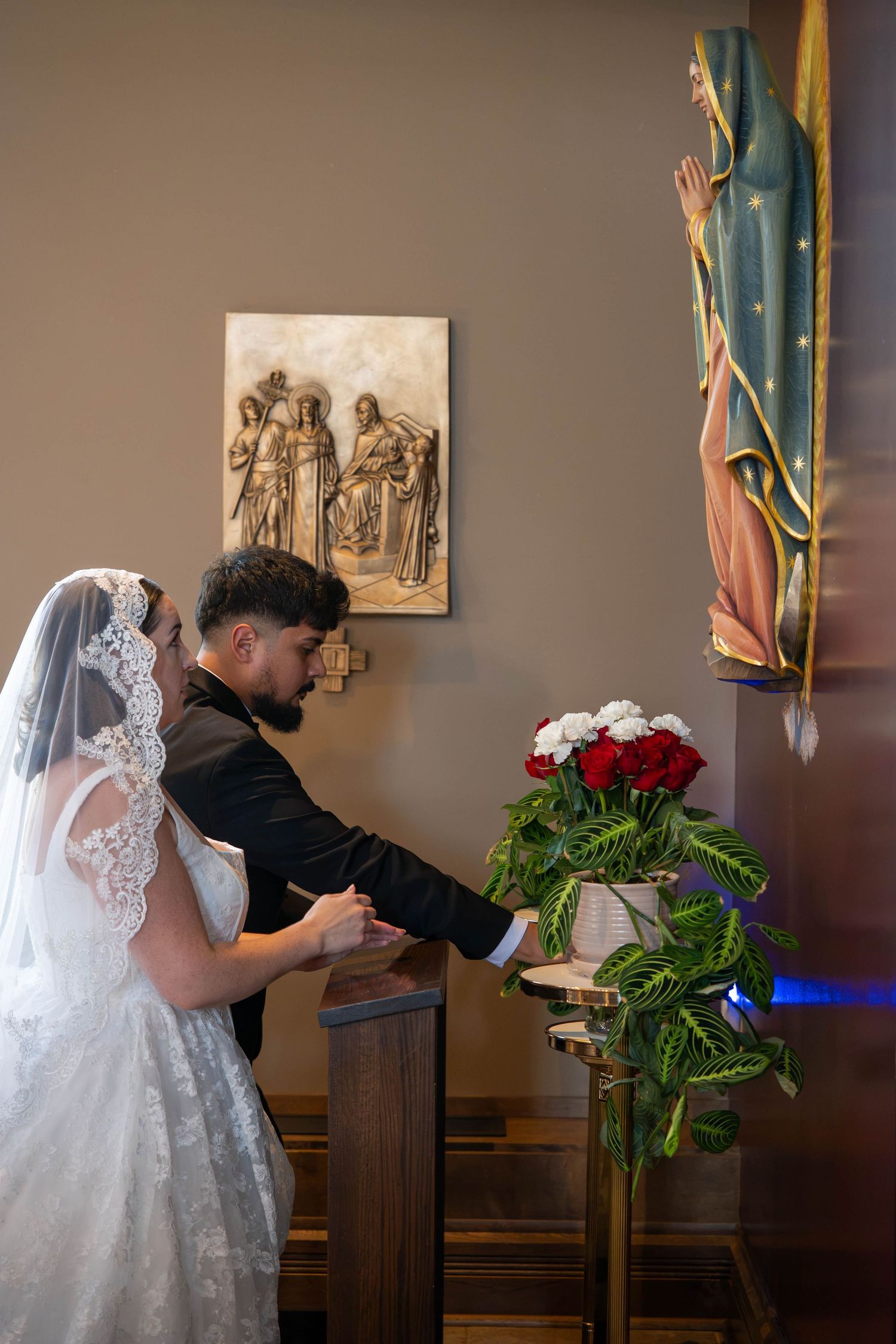 Bride and groom place flowers at Virgin Mary statue inside decorated Catholic church.