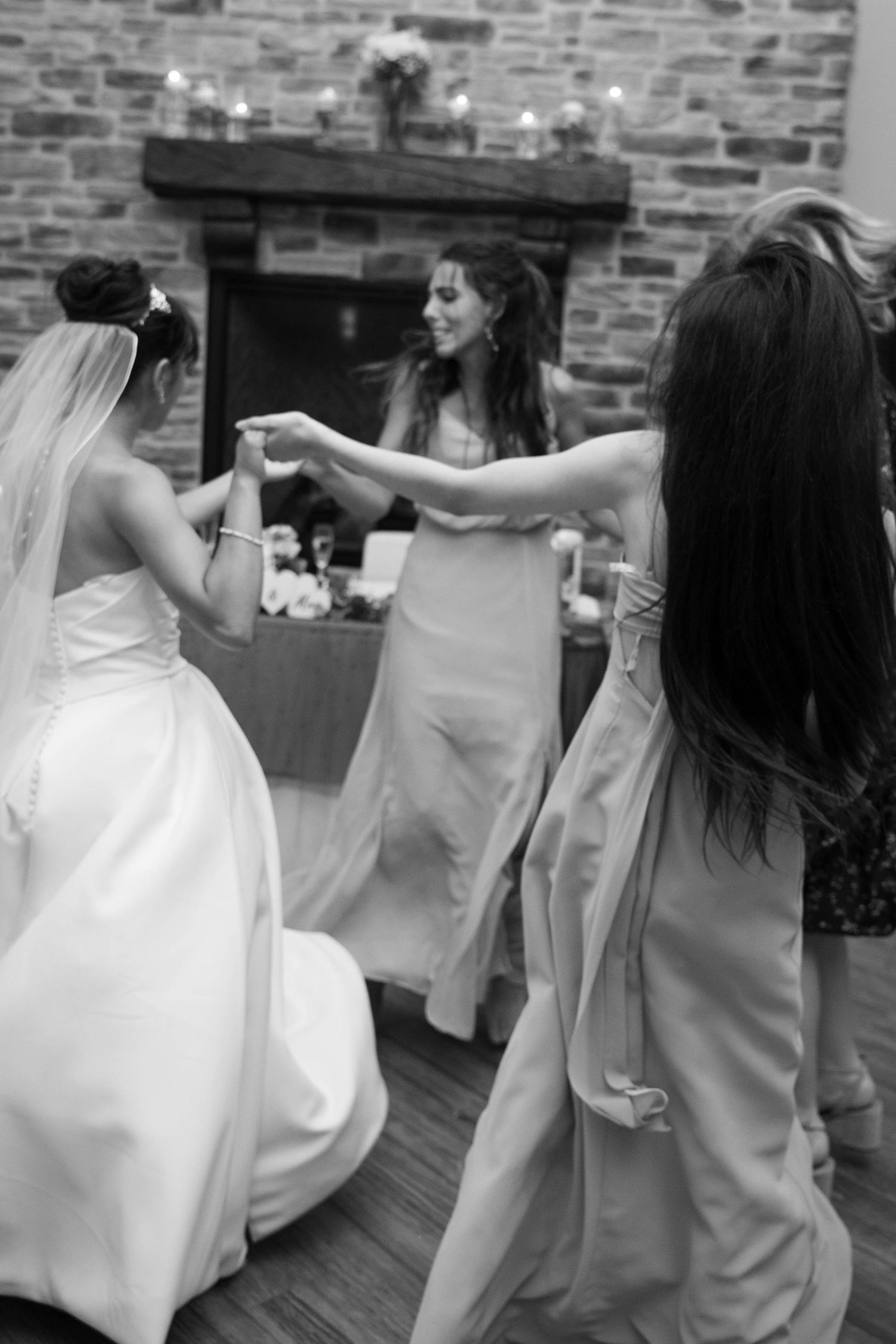 Black and white photo of bride dancing joyfully with bridesmaids near stone fireplace.