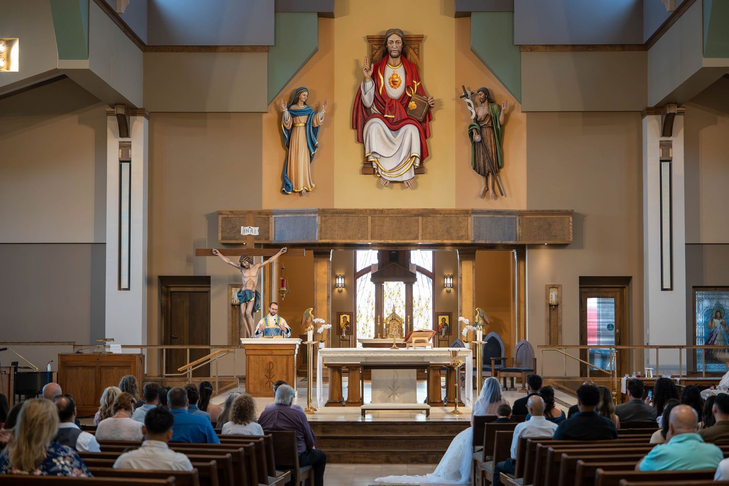 Wedding guests seated in wooden pews facing an ornate Catholic church altar with a large Jesus mural.