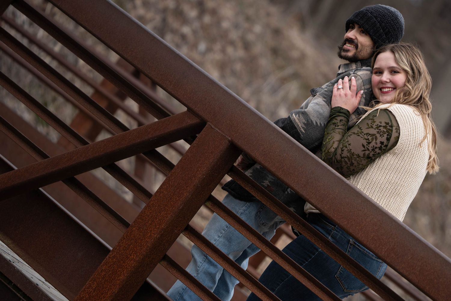 Couple embracing and laughing on rustic metal staircase railing outdoors in winter.