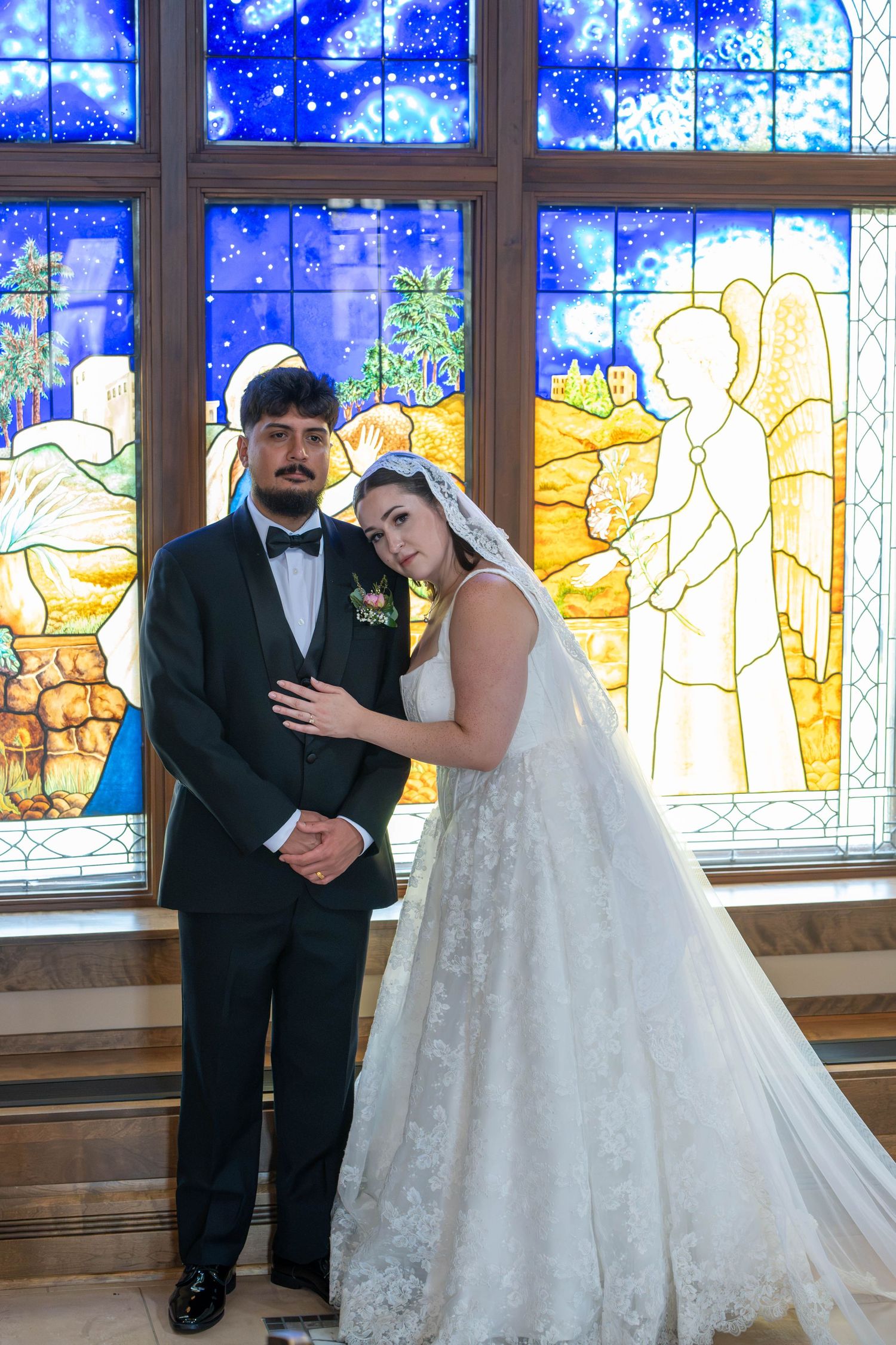 Bride and groom pose elegantly before vibrant stained glass window in church.