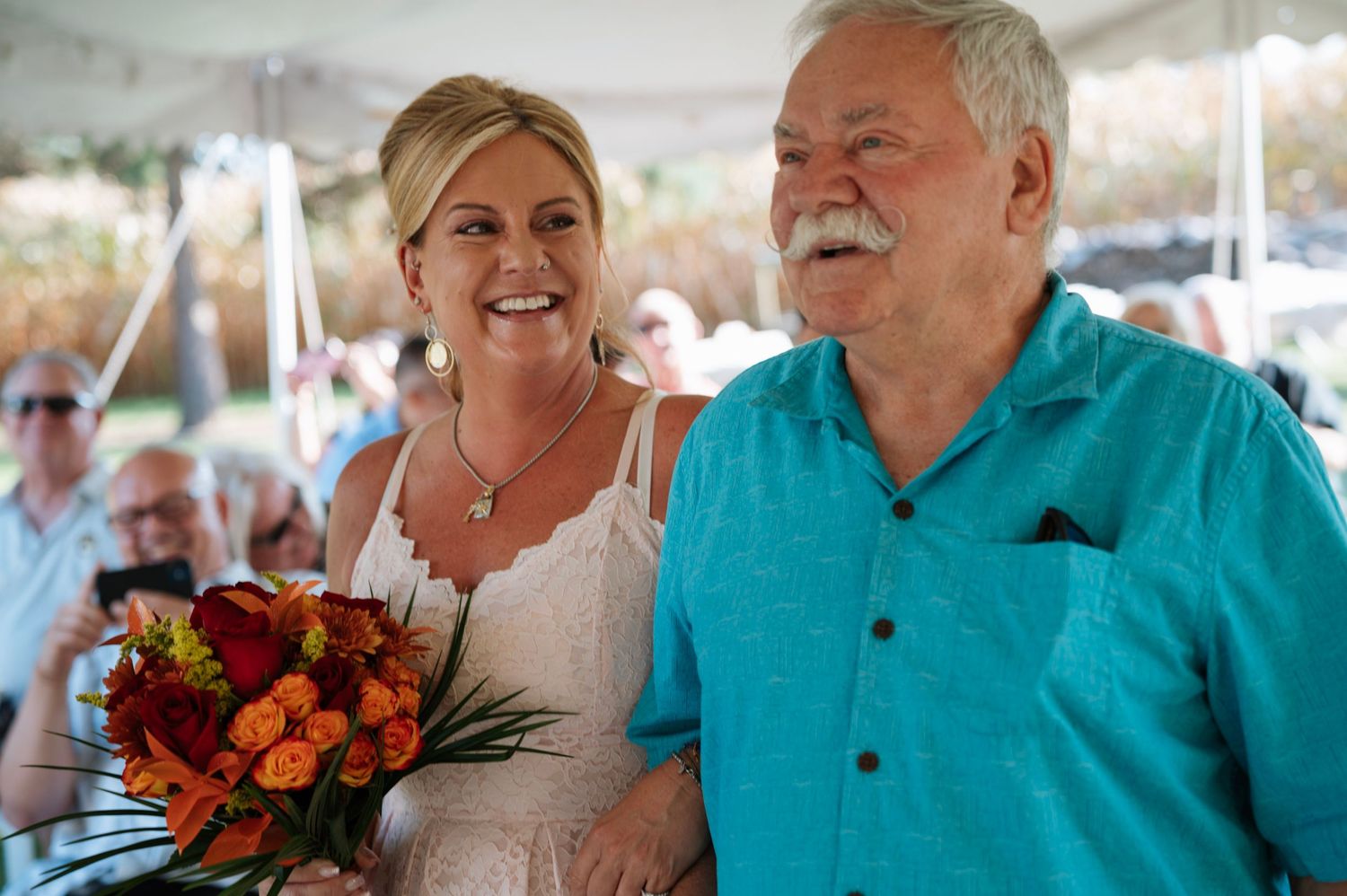 Smiling bride with autumn bouquet walking with older gentleman under wedding tent.