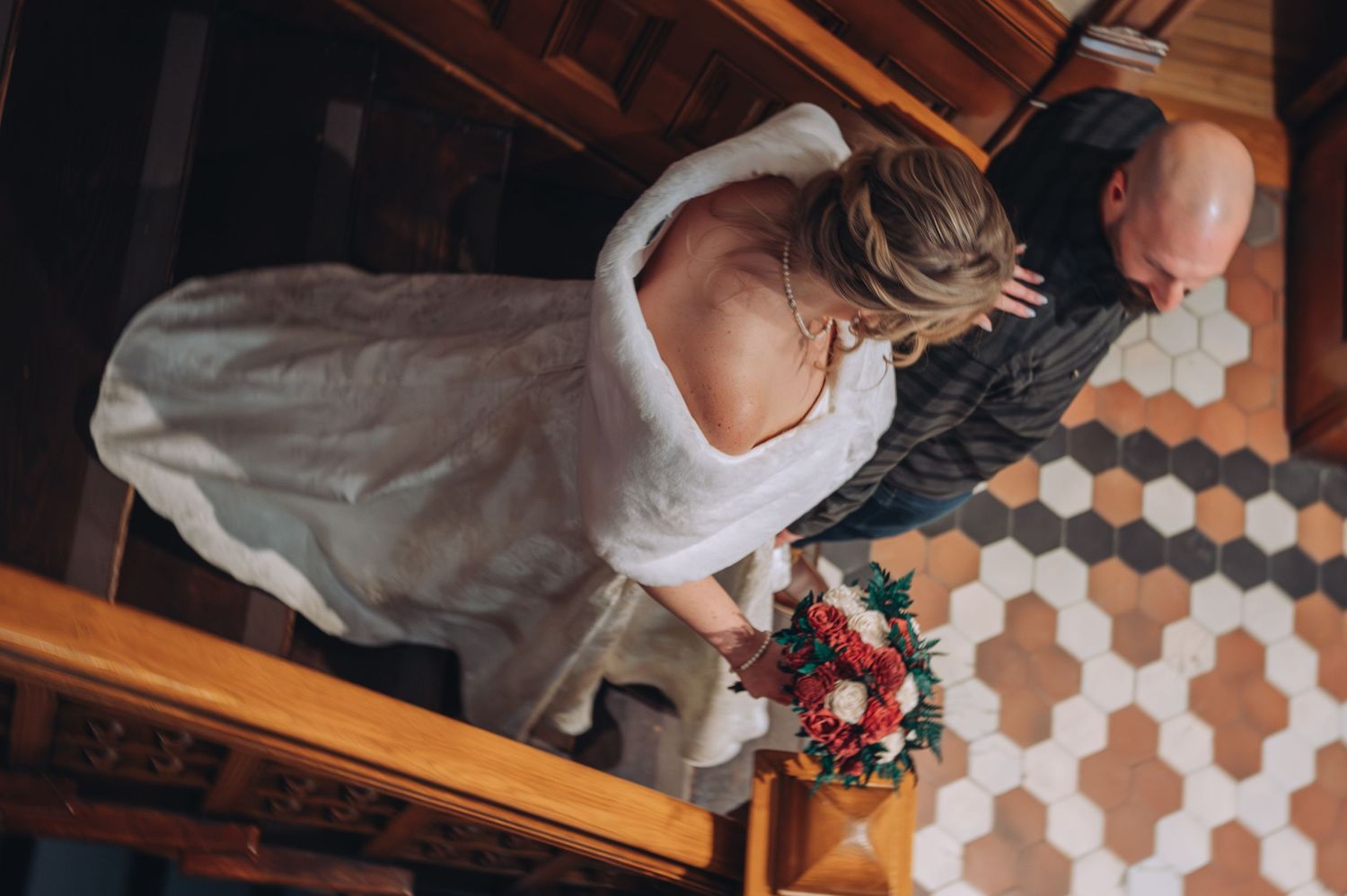 Aerial view of bride in white dress holding colorful bouquet walking with companion on hexagonal tile floor.