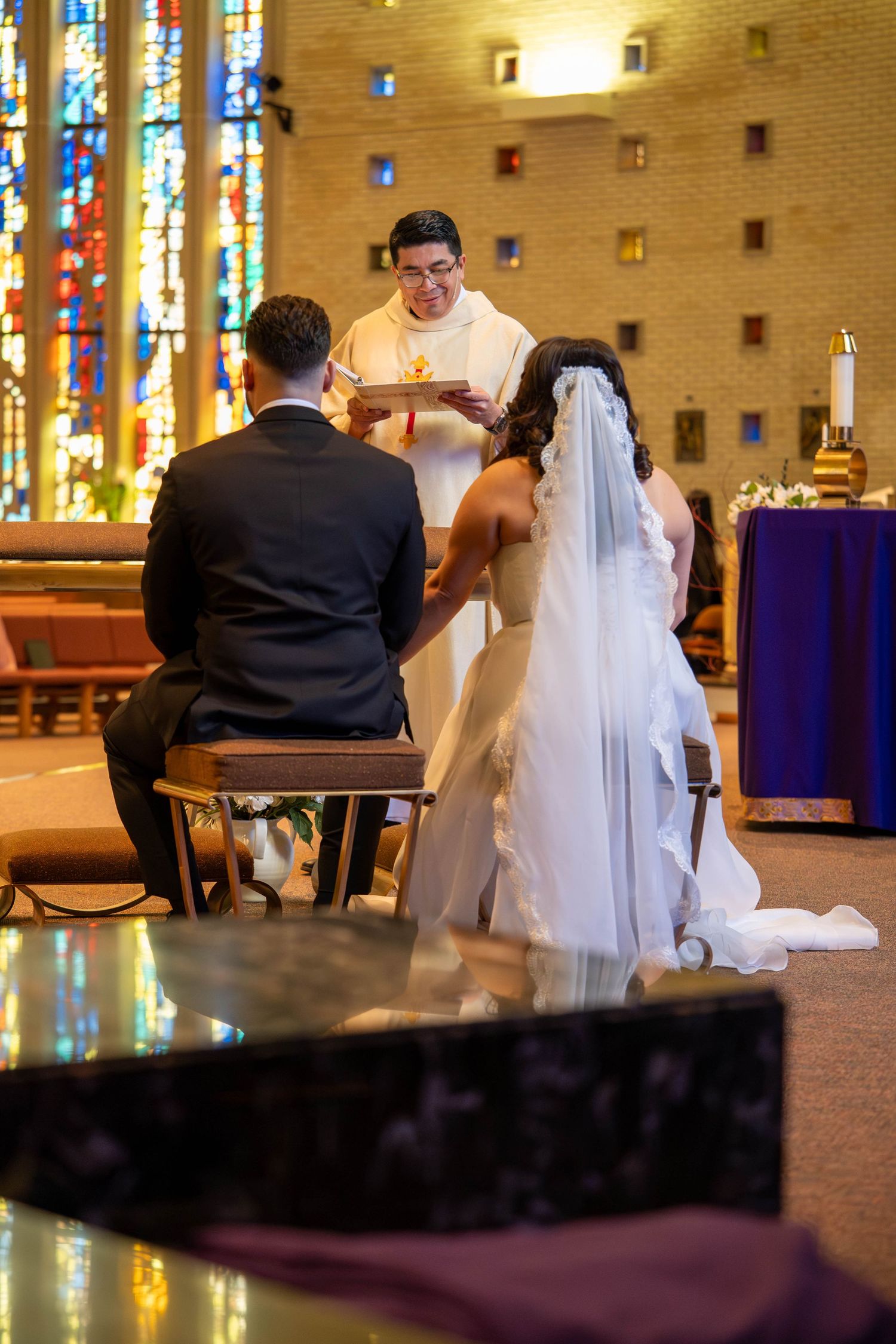 Bride and groom kneeling before priest during Catholic wedding ceremony with stained glass windows in background.