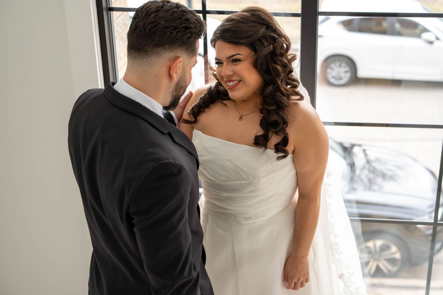 Joyful bride in white strapless gown smiles at groom in black suit by window.