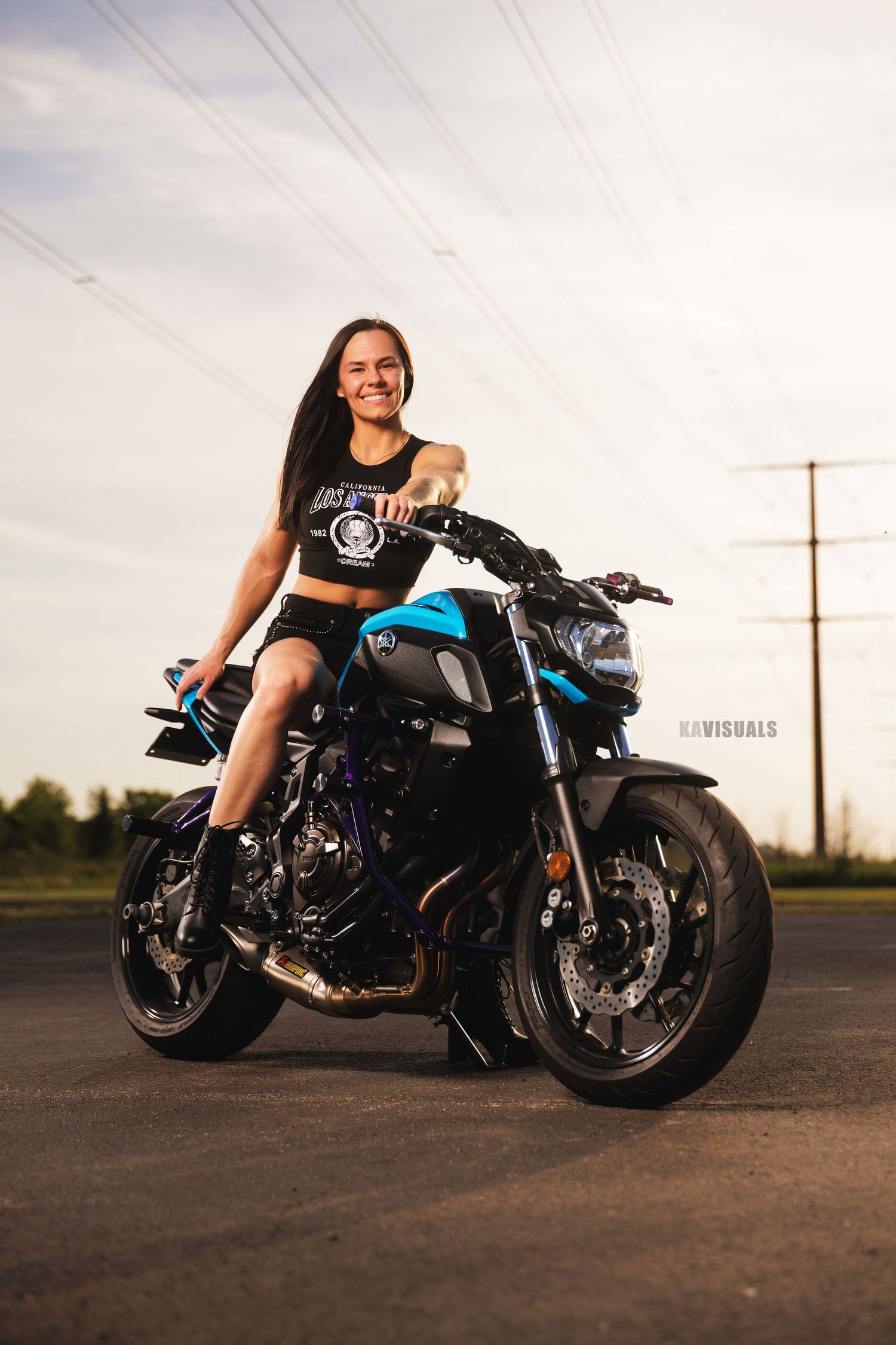 Smiling athletic woman sitting on blue Yamaha MT-07 motorcycle outdoors at sunset.