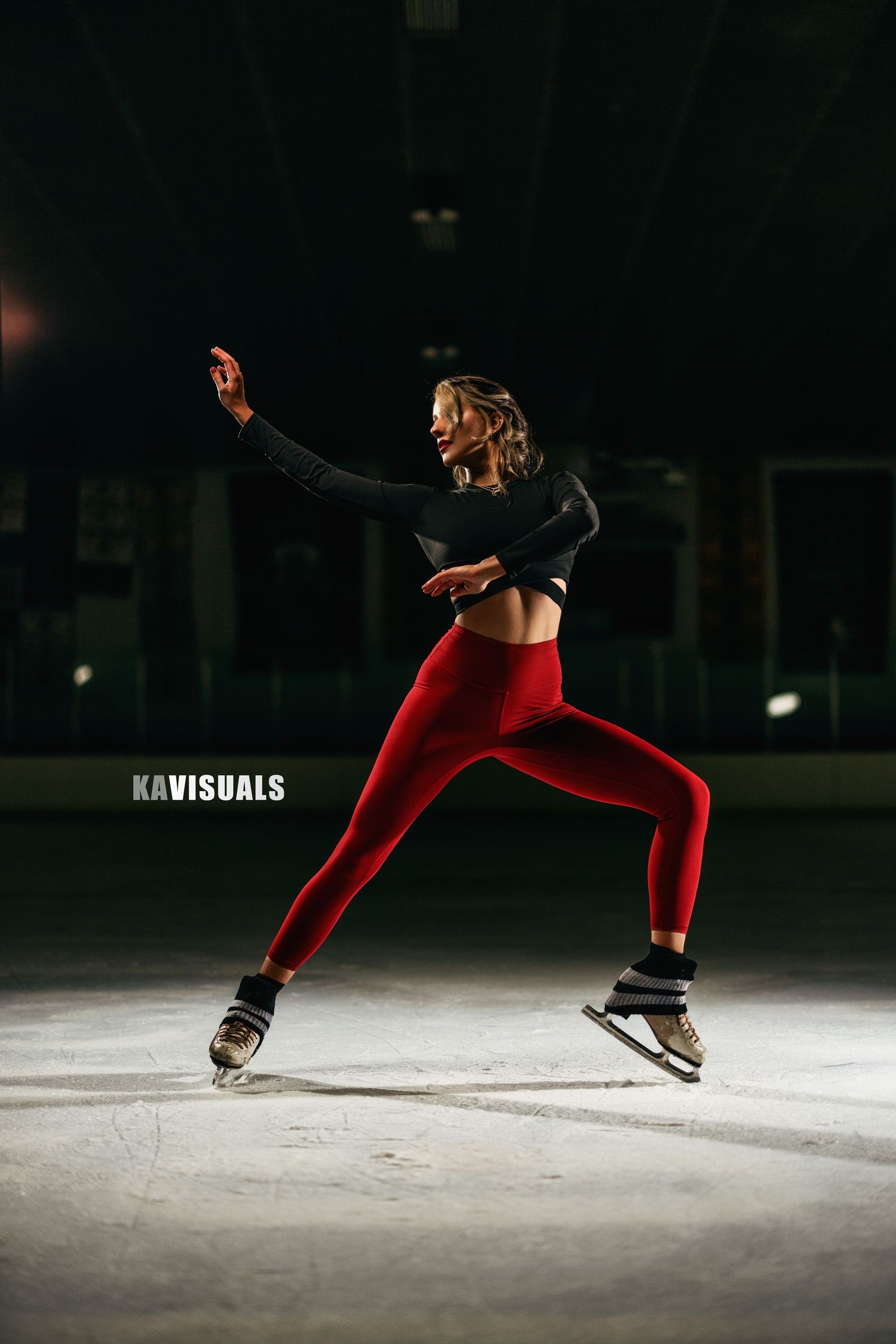 Female figure skater in red leggings and black top executing a graceful skating pose on an indoor ice rink.