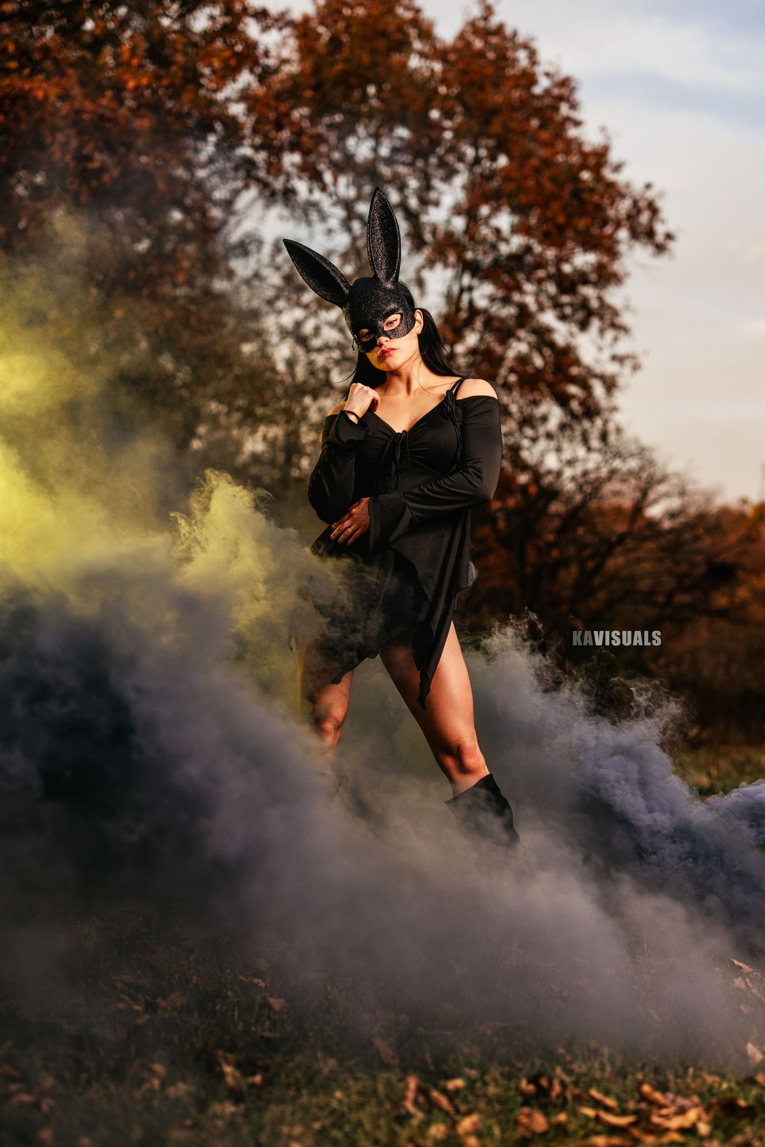 Woman in black dress and bunny mask surrounded by colorful smoke in autumn park.