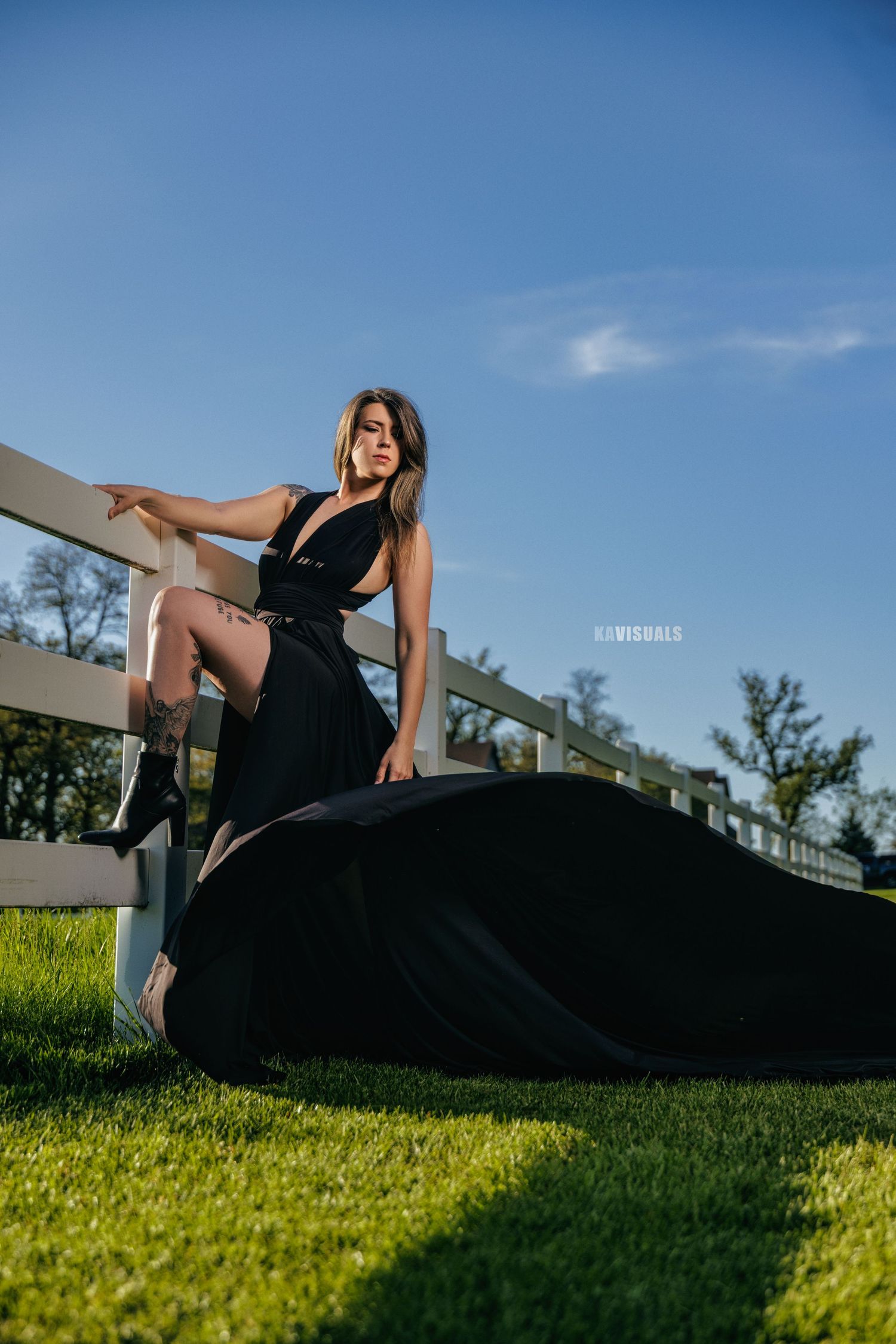 Woman in black dress posing by white fence on green grass.