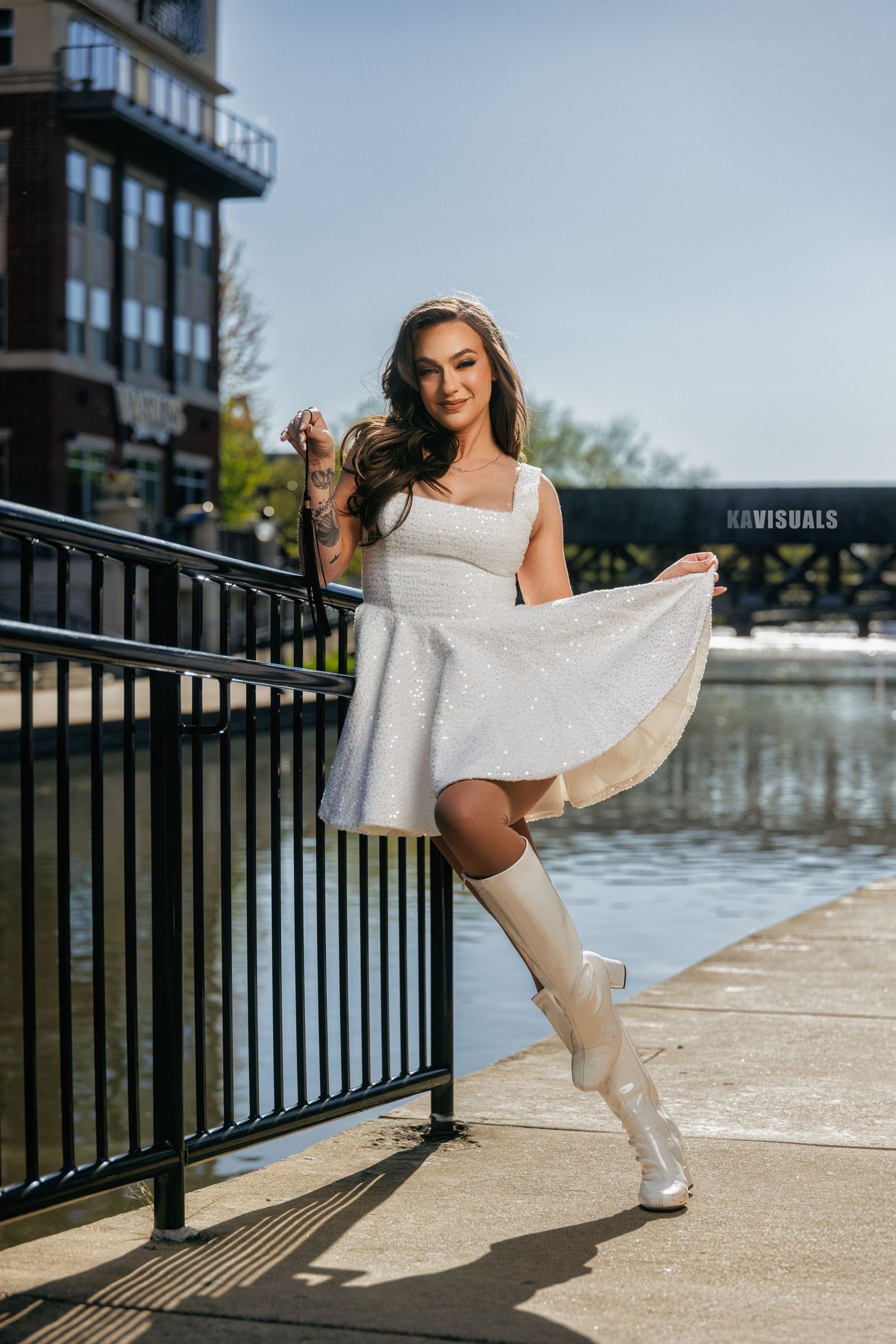 Brunette woman in a white mini dress and knee-high boots posing by a railing near a waterway.