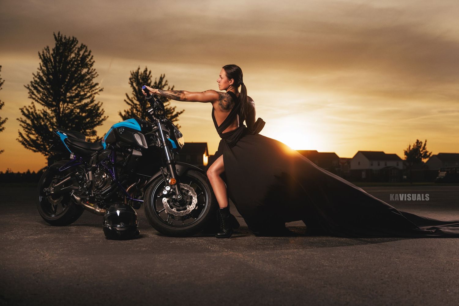 Woman in black outfit leaning against blue motorcycle during dramatic golden sunset outdoors.
