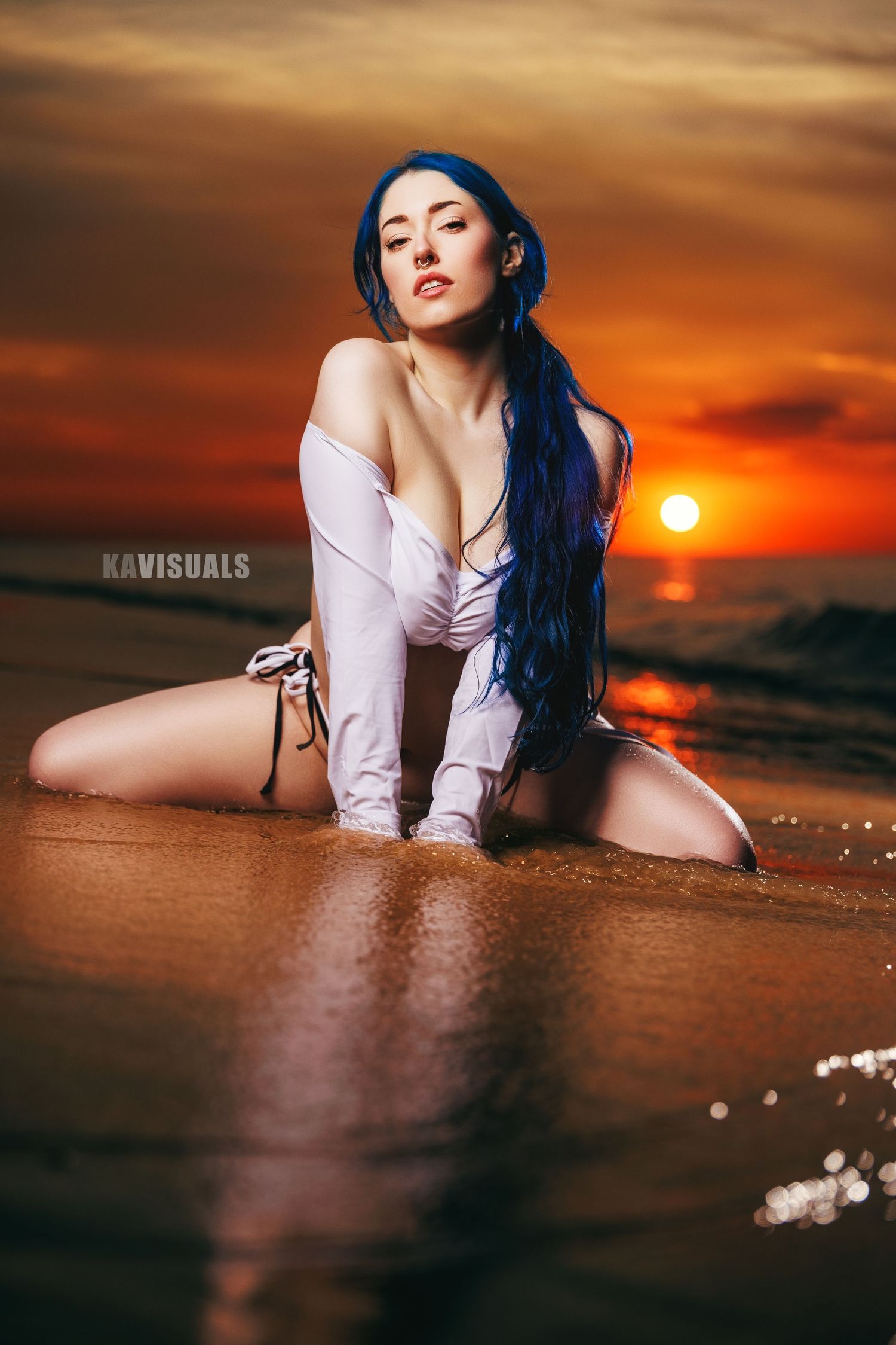 Woman with blue hair in white swimsuit poses on beach at sunset with ocean waves.