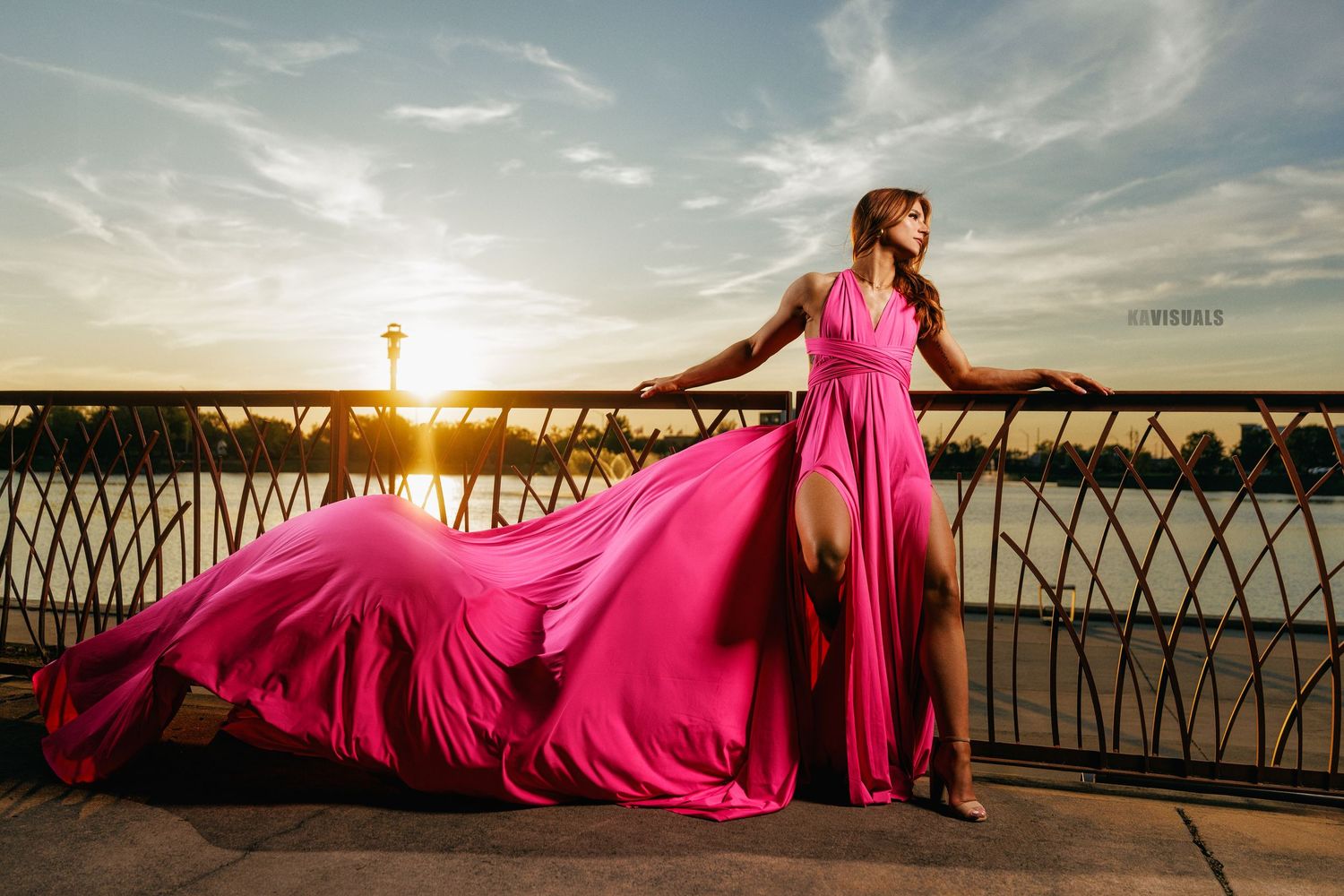 Woman in flowing pink gown posing on bridge at golden hour sunset.