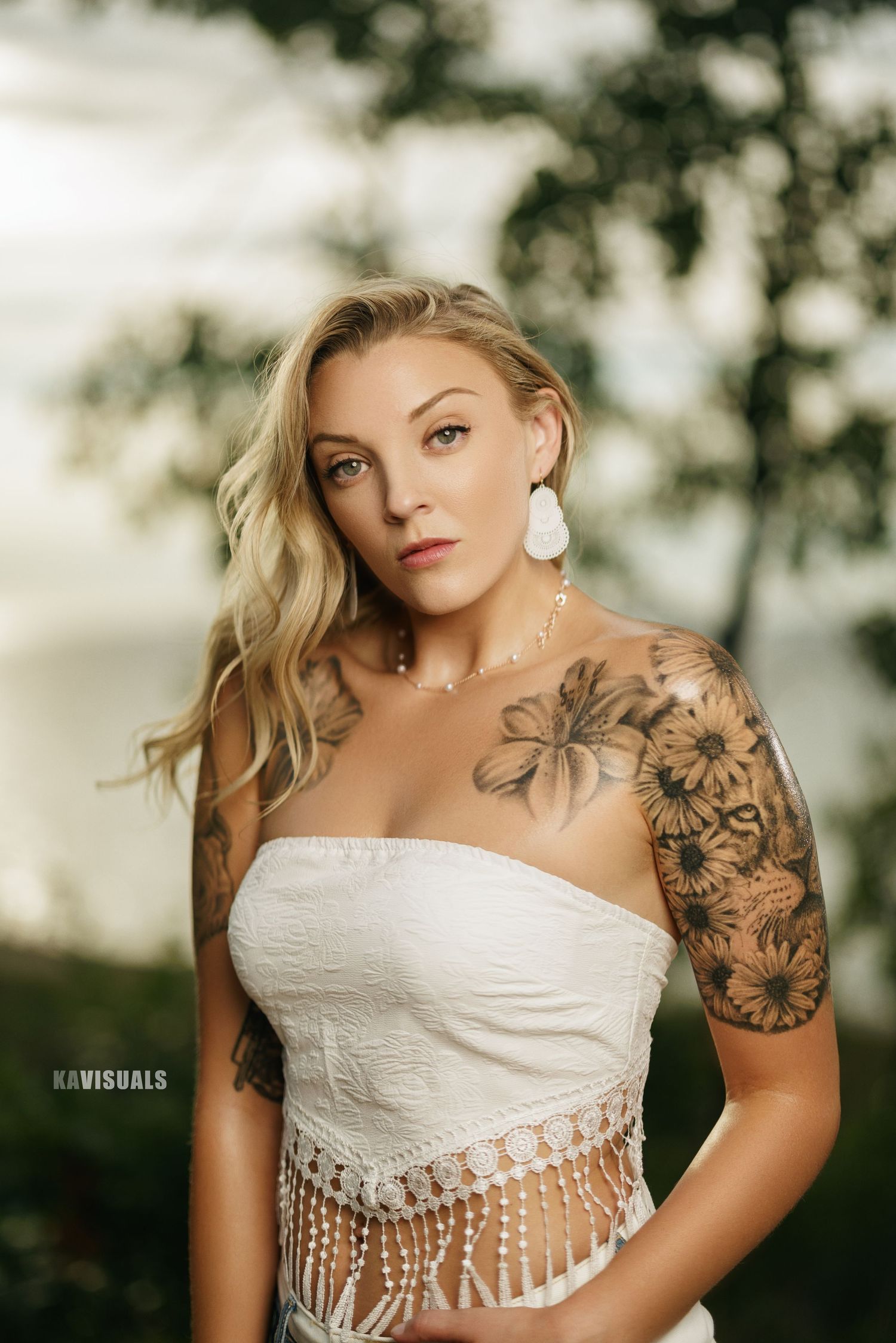 Tattooed blonde woman in white fringe strapless top posing outdoors near trees with soft misty background.