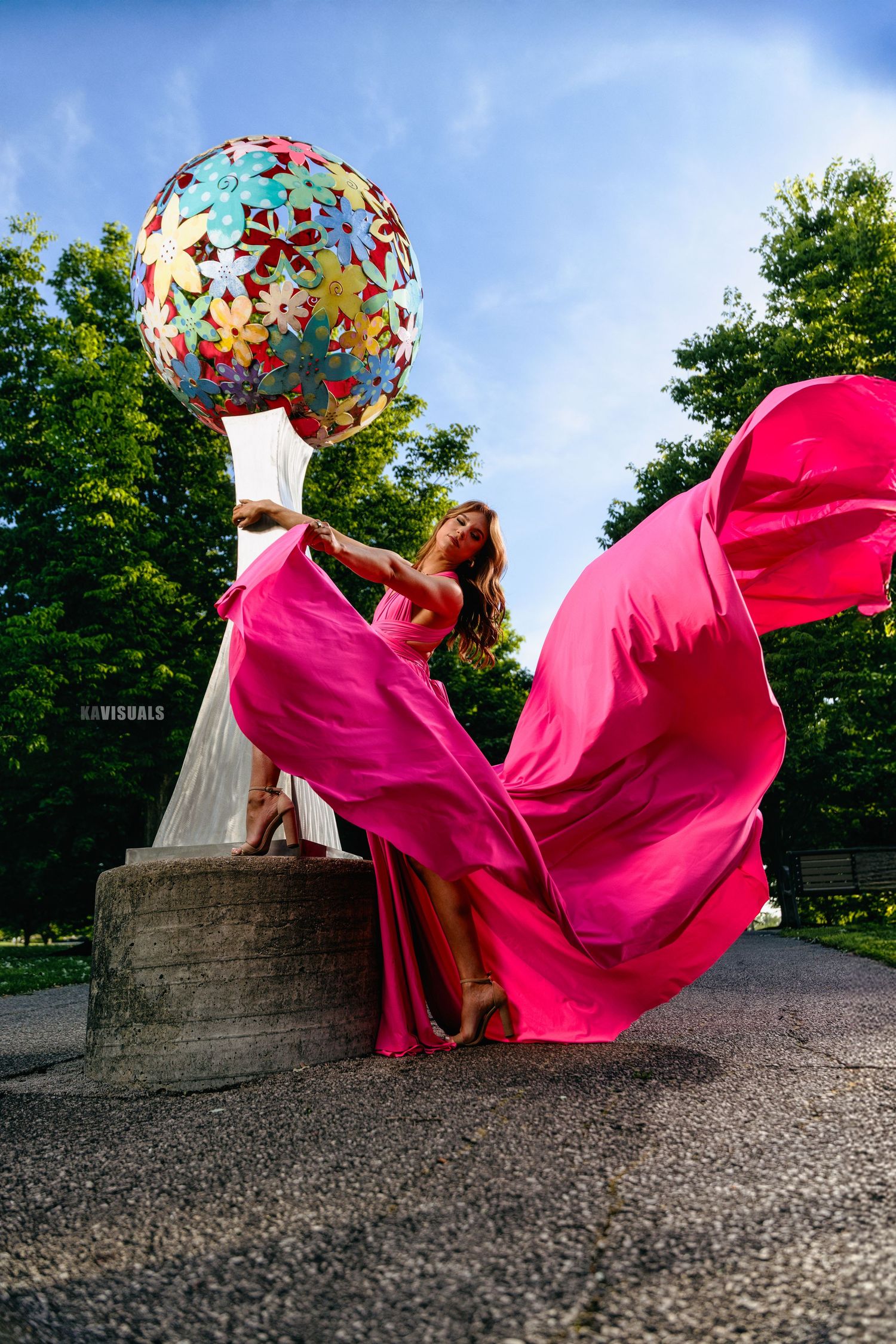 Woman in flowing pink dress dancing near colorful floral sculpture in park.
