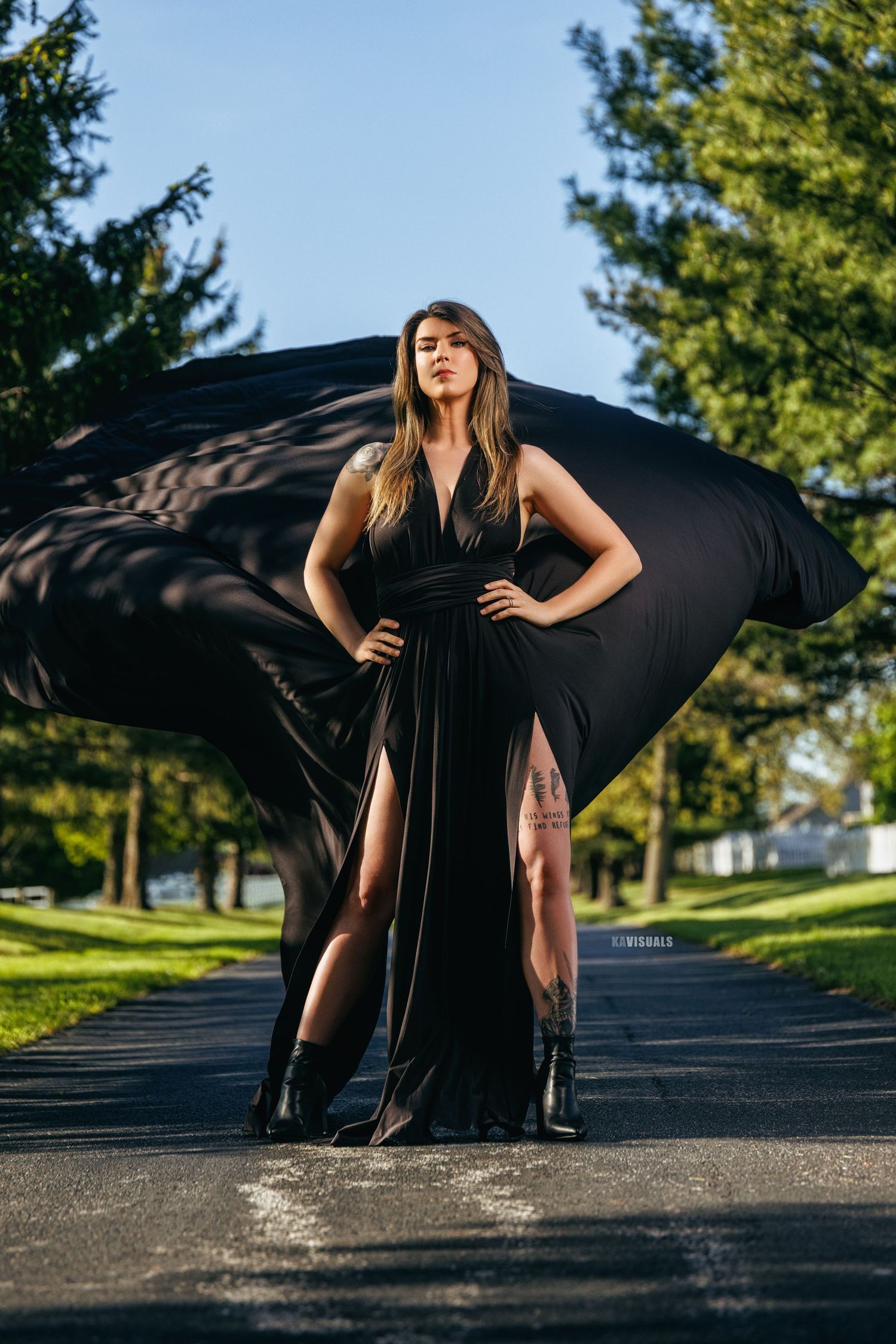 Woman in black gown with billowing fabric on tree-lined road.