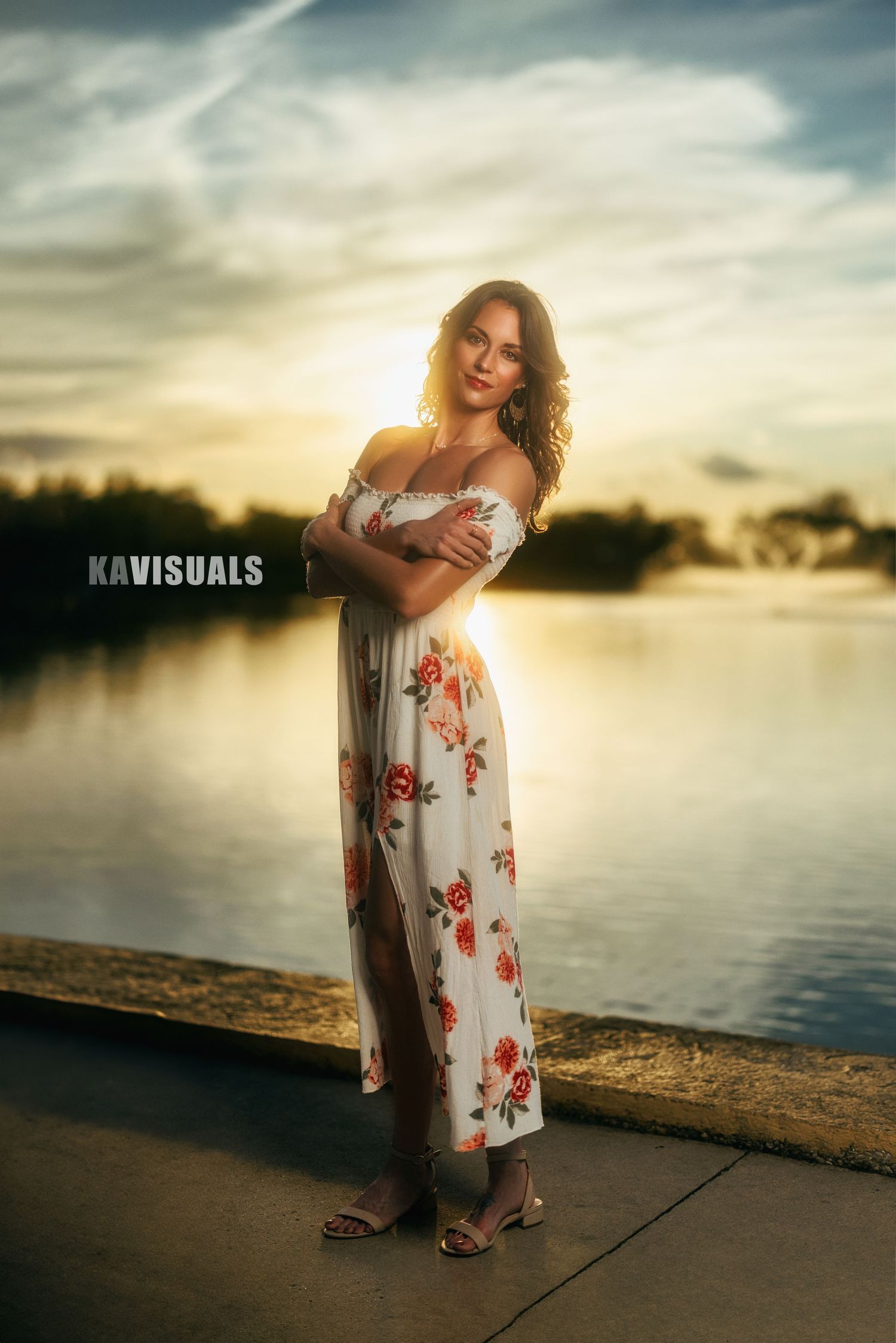 Woman in floral off-shoulder maxi dress poses by lake during golden sunset.