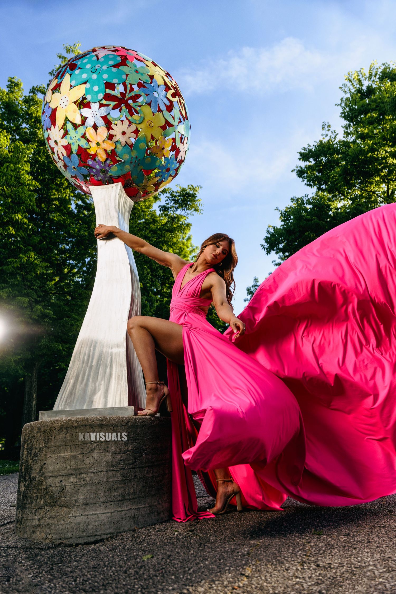Woman in a vibrant pink flowing dress posing beside a colorful floral sphere sculpture outdoors.