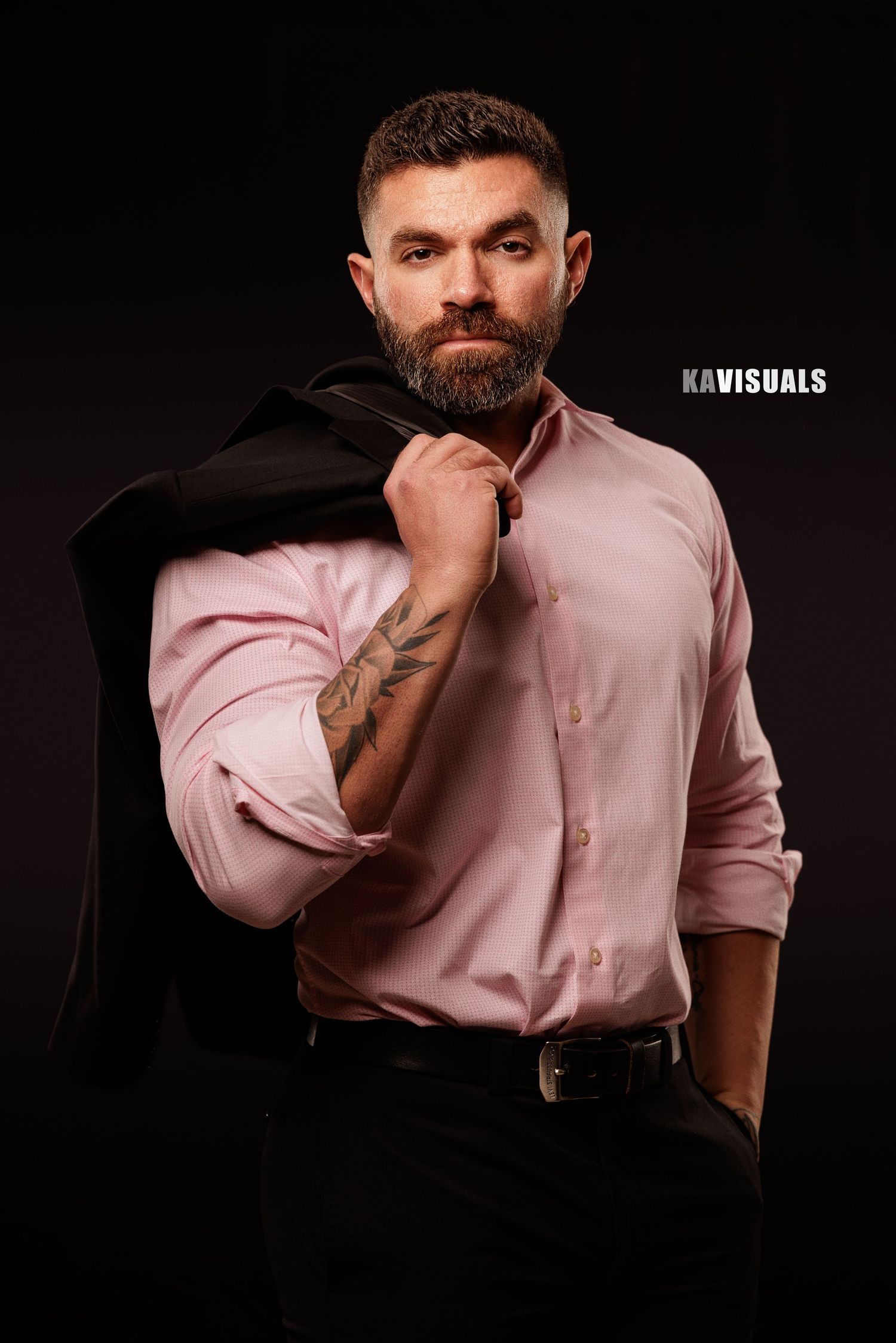 Bearded man in pink shirt holds jacket over shoulder against dark background.