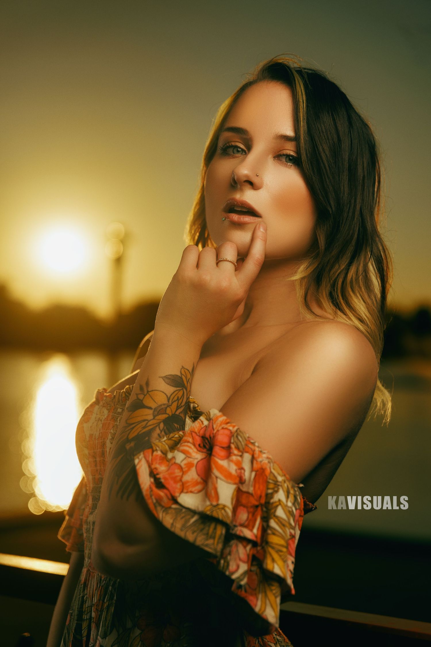 Woman with tattoos in floral top poses at waterfront during sunset.