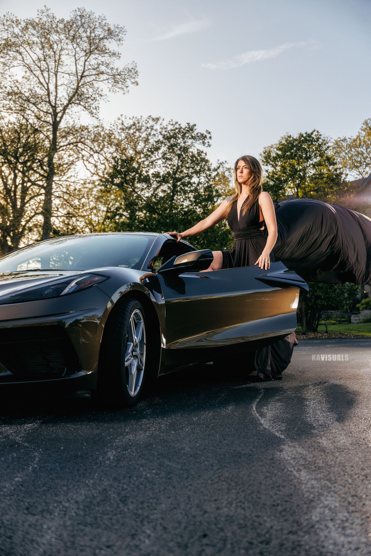 Woman in black dress unveiling a sleek black sports car outdoors at golden hour.